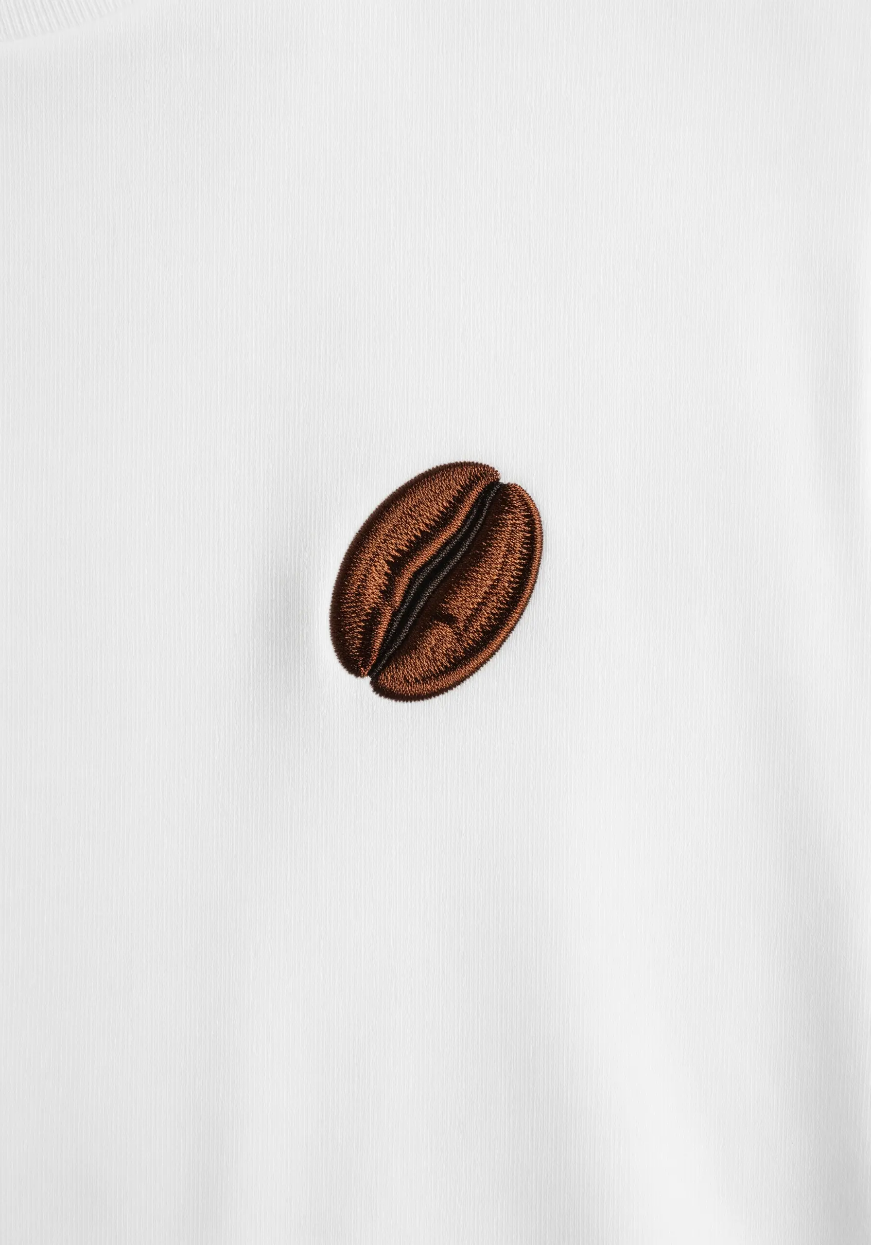 A realistic coffee bean embroidered with brown satin stitch on a white shirt.