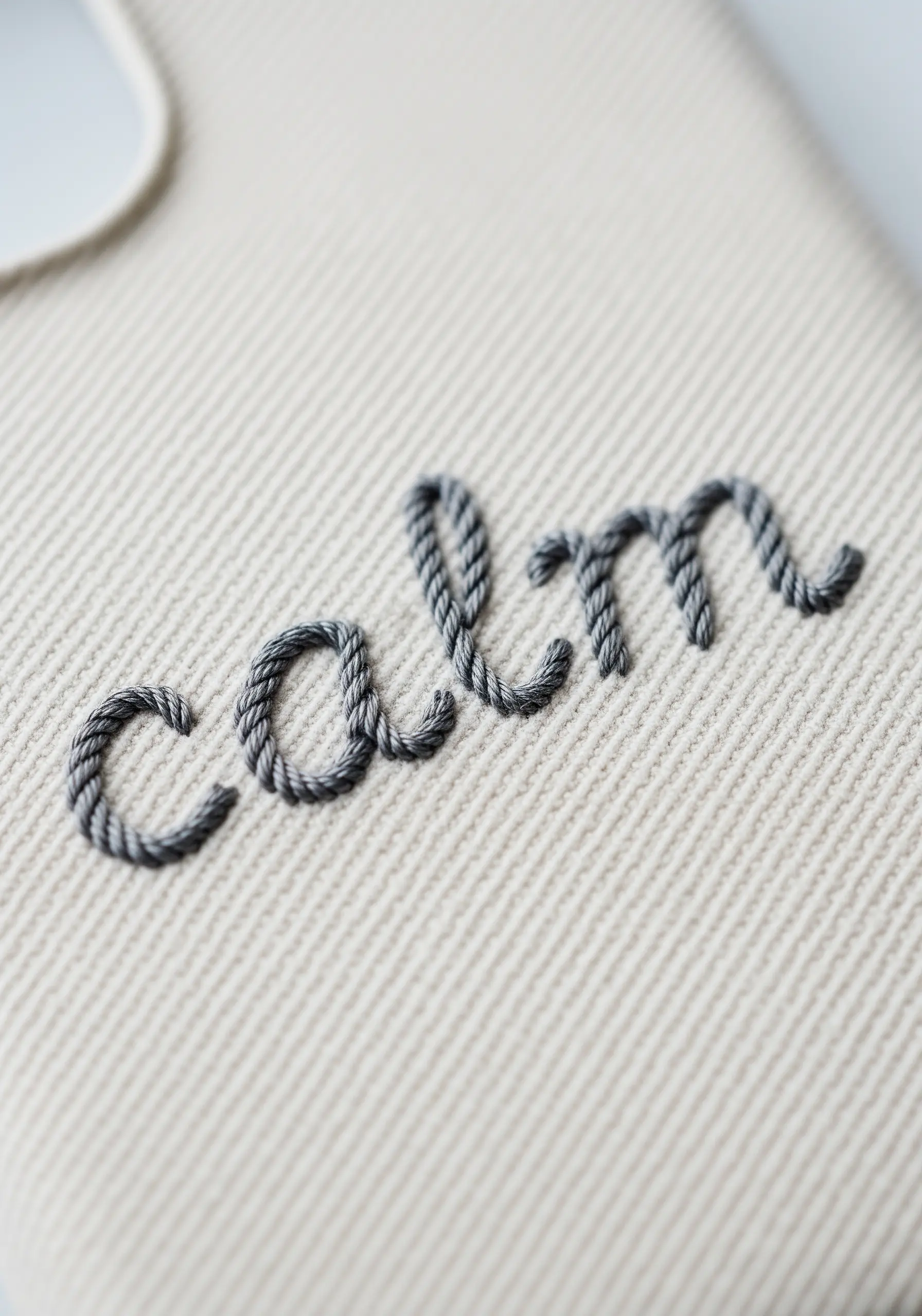 The word 'calm' embroidered in a raised, rope-like stitch using grey thread on a cream case.