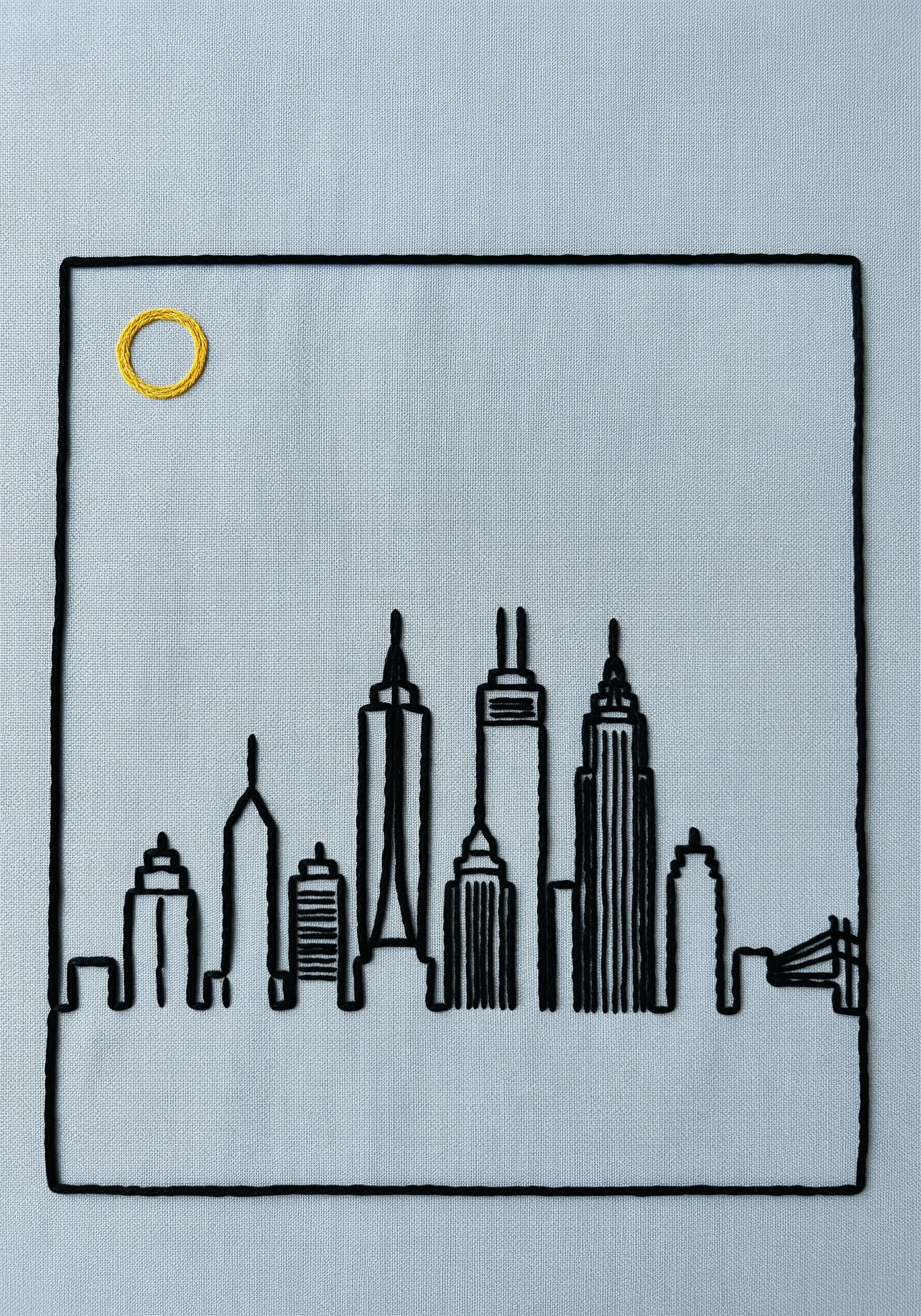 Black thread outline of a city skyline with a small, bright yellow embroidered sun.
