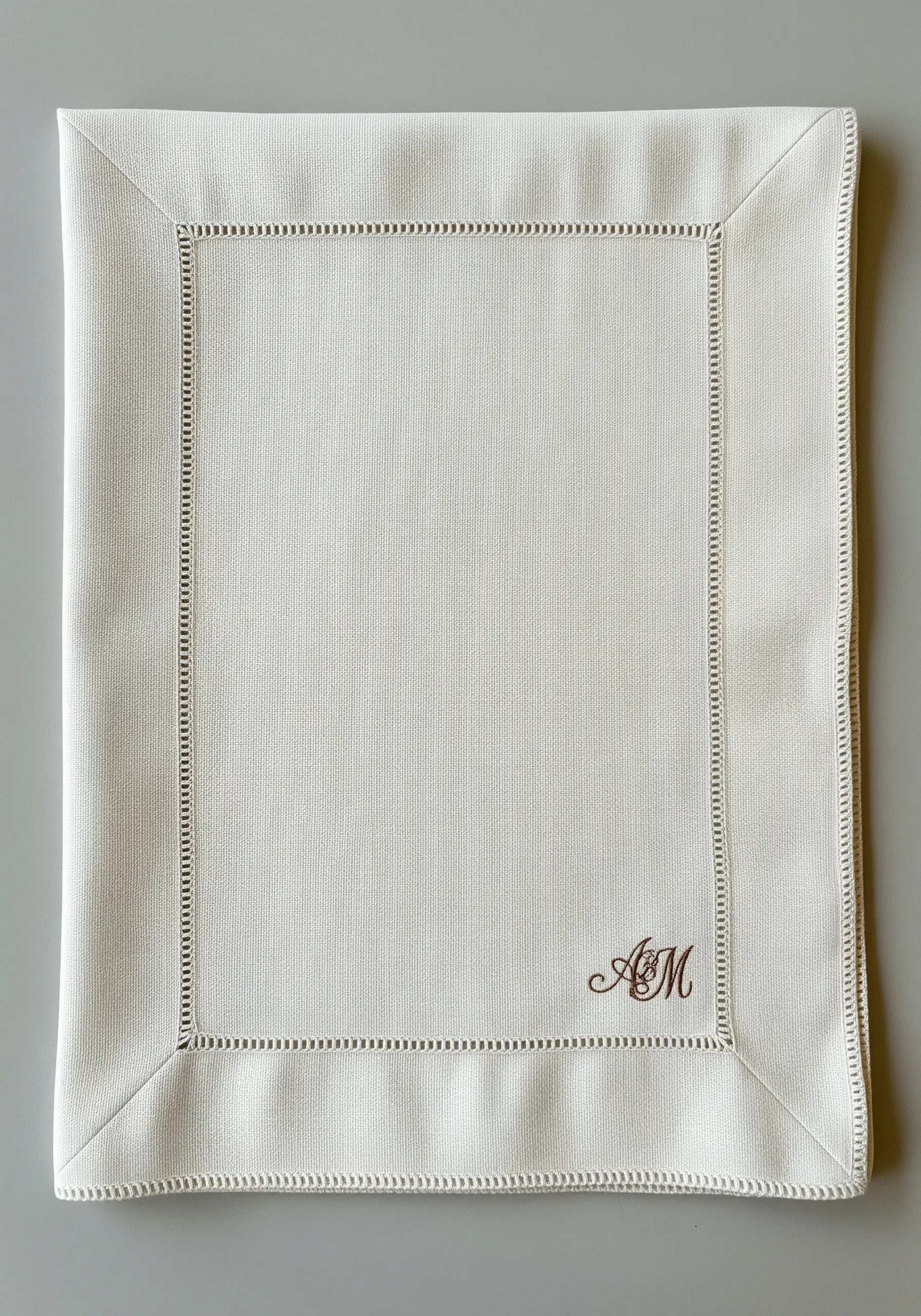 A small, elegant 'AM' monogram stitched in brown thread on a cream linen placemat.