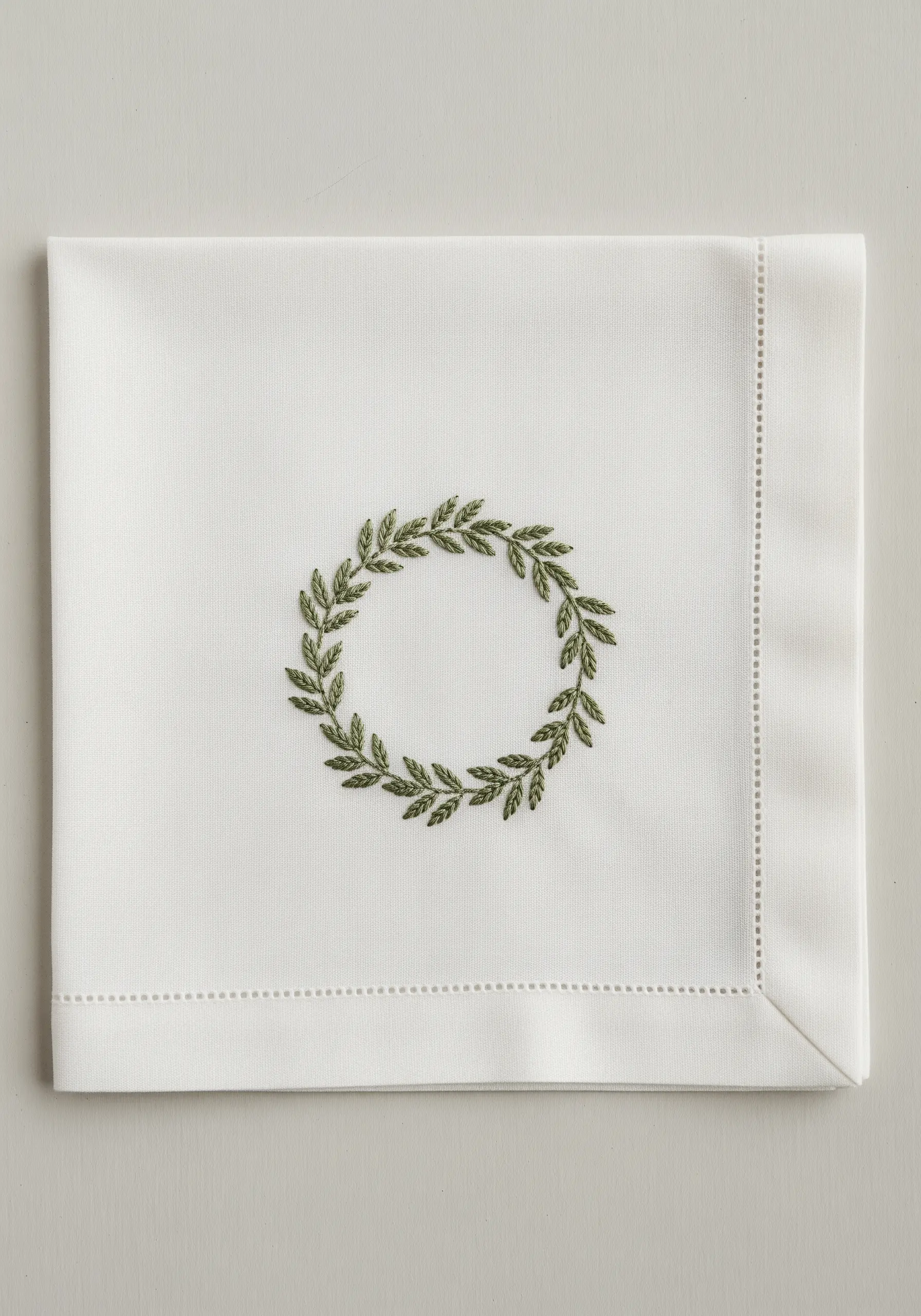 A minimalist olive leaf wreath embroidered in green on a hemstitched white napkin.