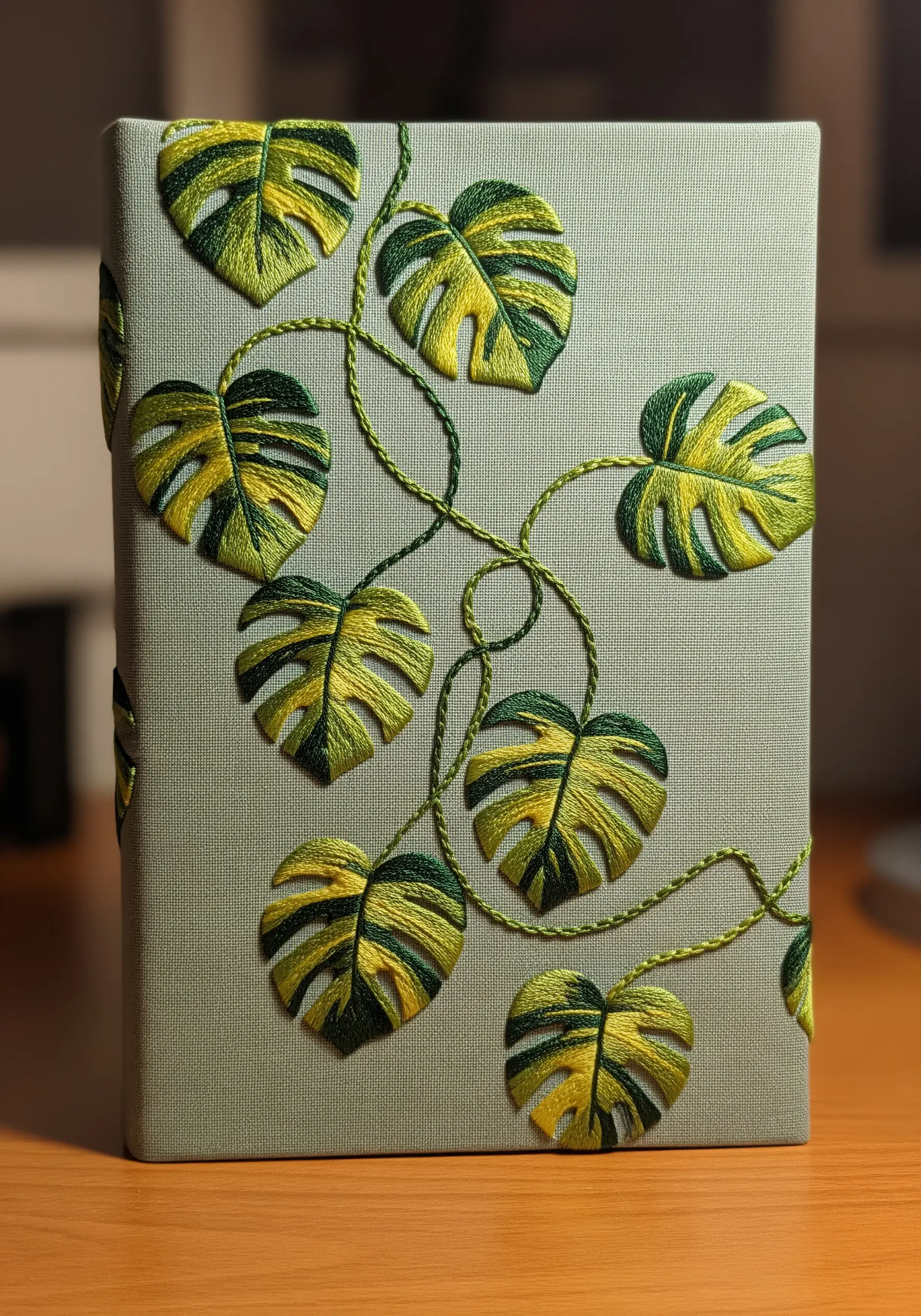 Multiple small, variegated Monstera leaves embroidered onto a sage green book cover.