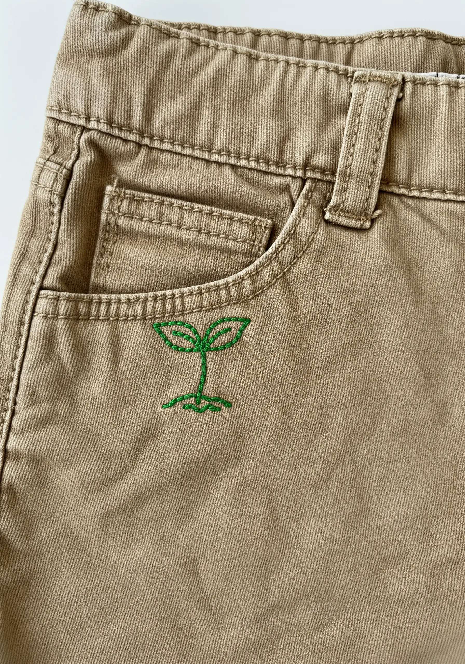 Green embroidered sprout icon stitched near the pocket of khaki pants.