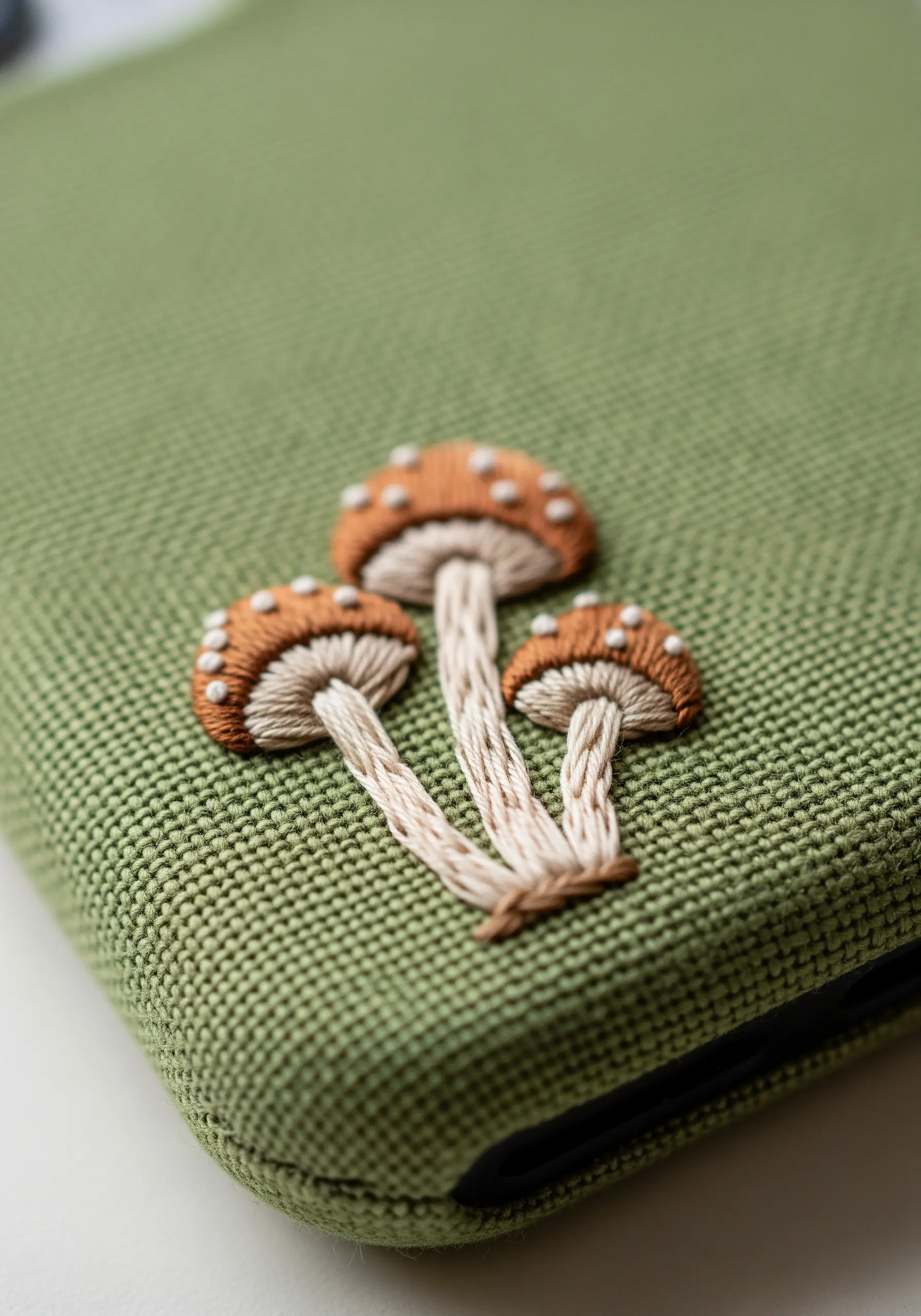 Three small, detailed mushrooms embroidered in brown and white on a green phone case.