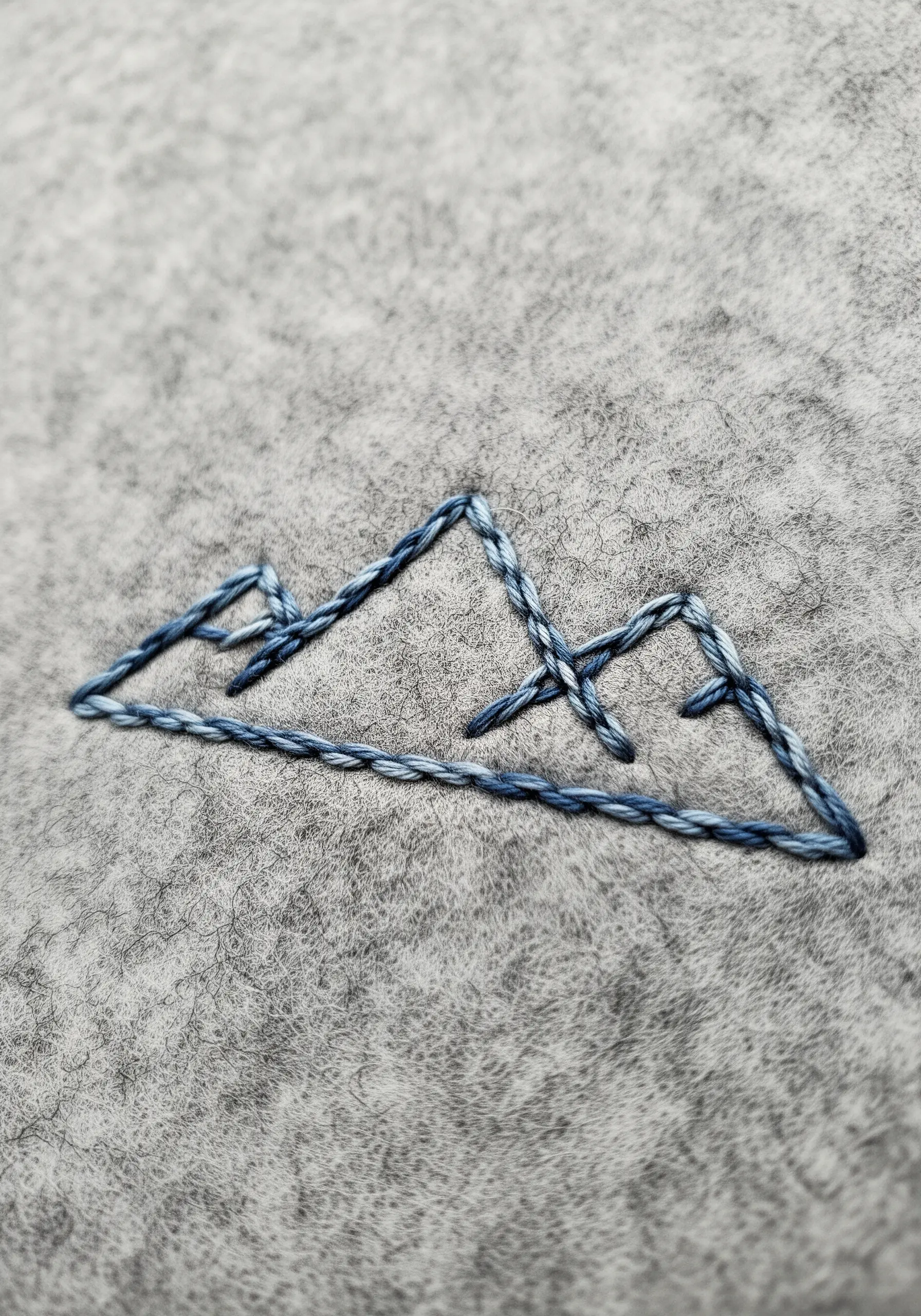 A minimalist mountain range outline stitched with blue variegated thread on grey felt.