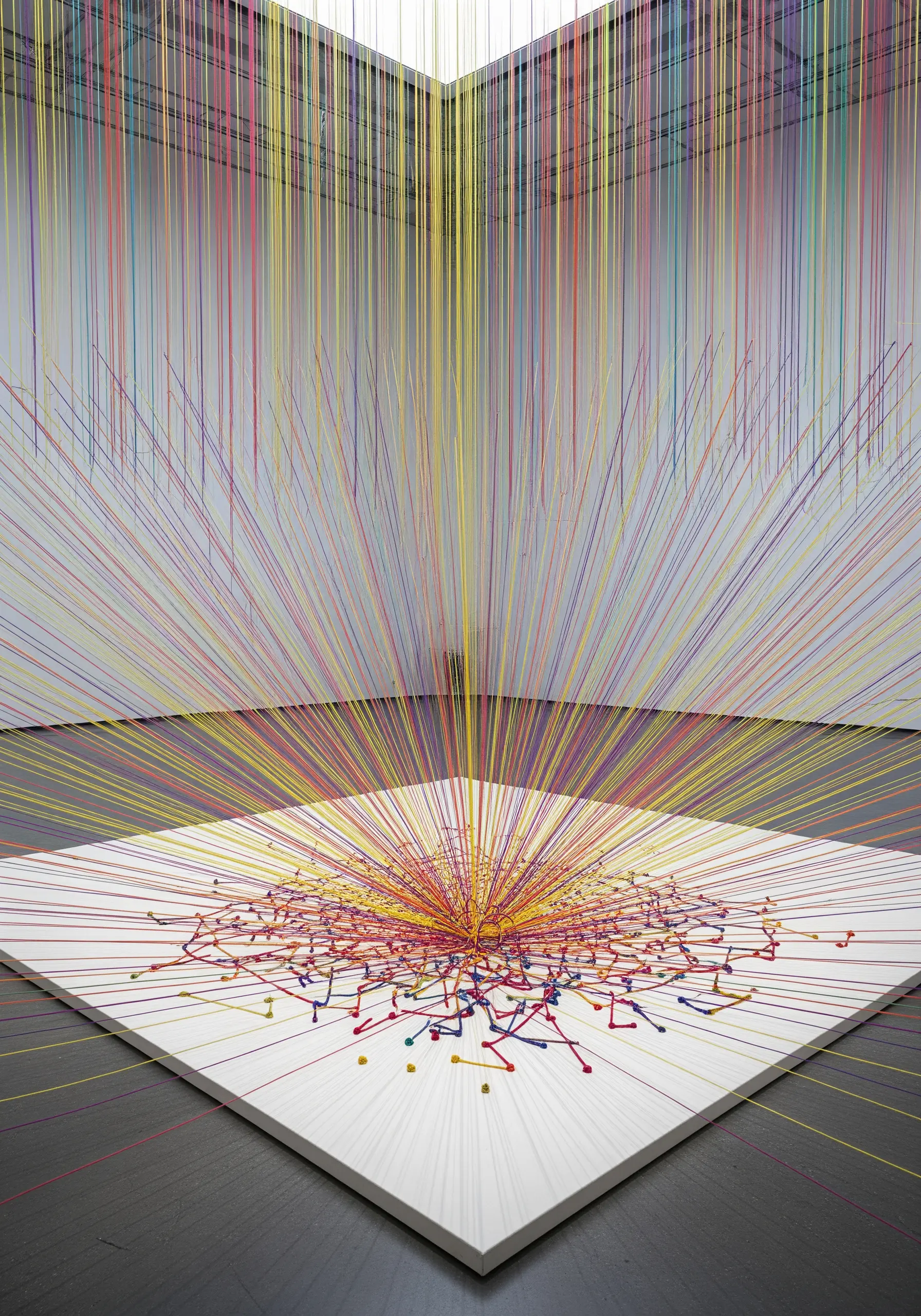 A large-scale room installation with colorful threads stretching from a point on the floor to the ceiling.