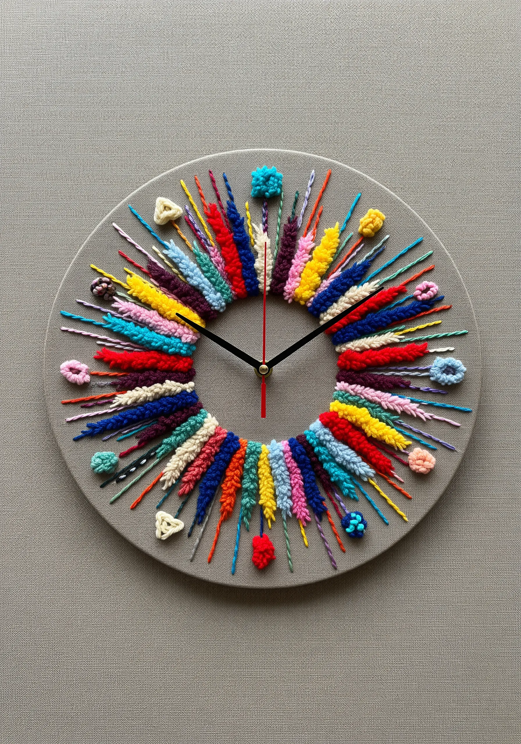 A functional wall clock with a gray fabric face, embroidered with colorful, textural stitches for hours.