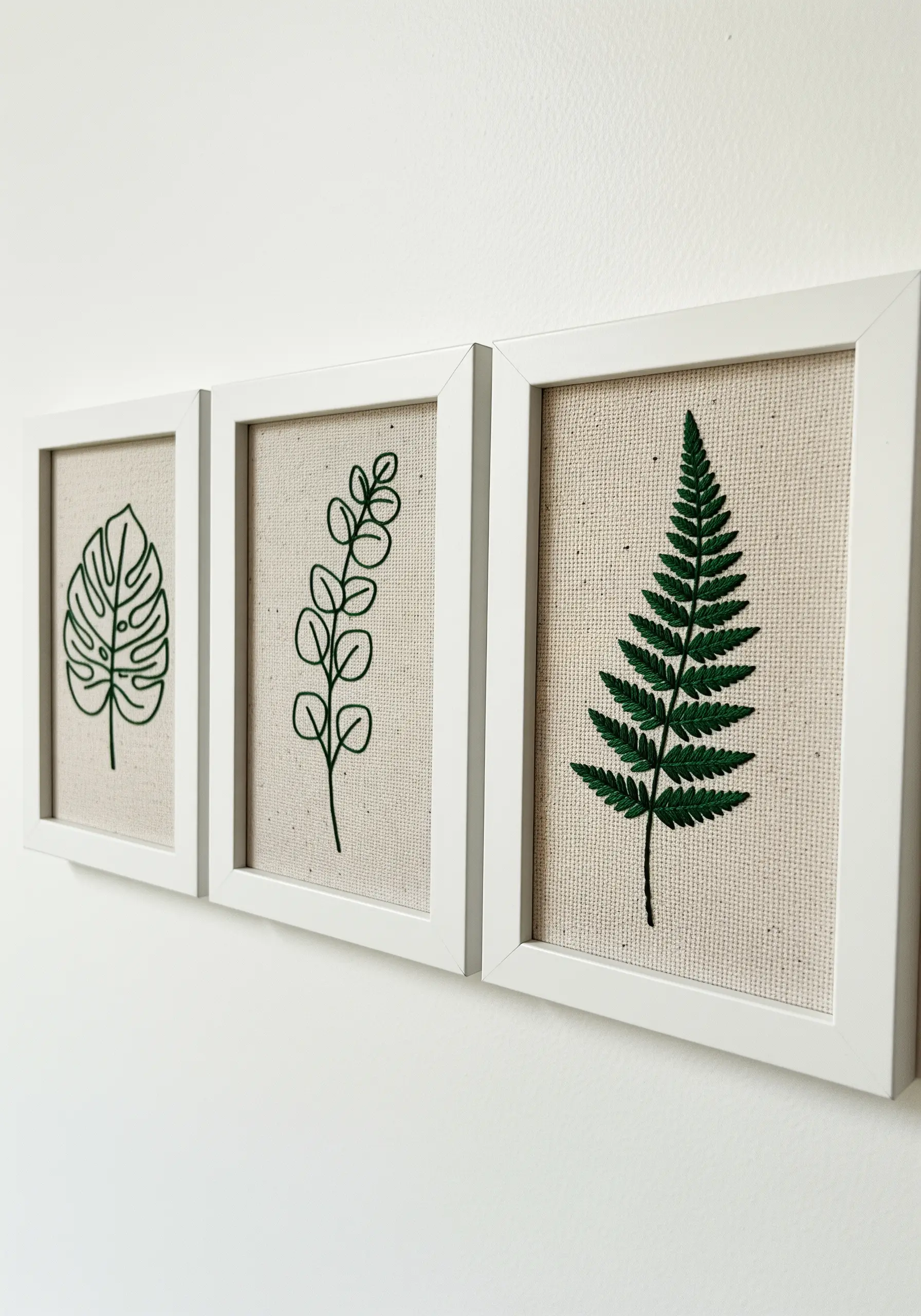 A triptych of three framed botanical embroideries: monstera, eucalyptus, and fern.