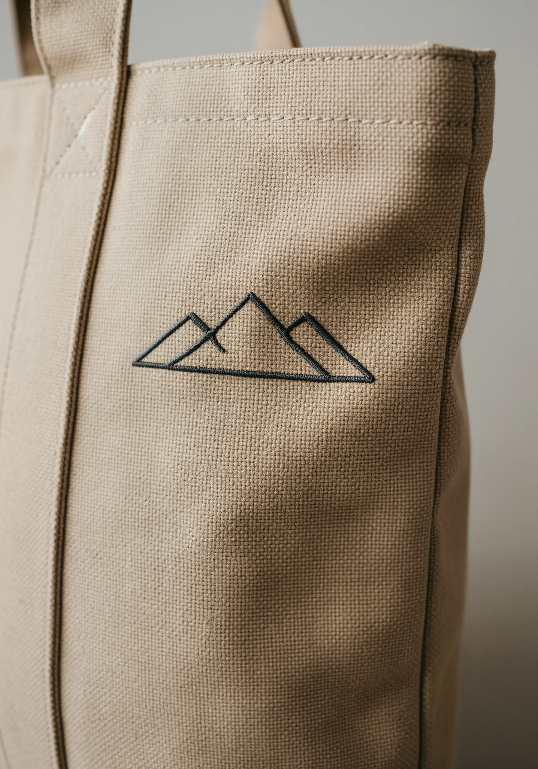 A minimalist mountain range logo stitched in black thread on a beige canvas tote bag.