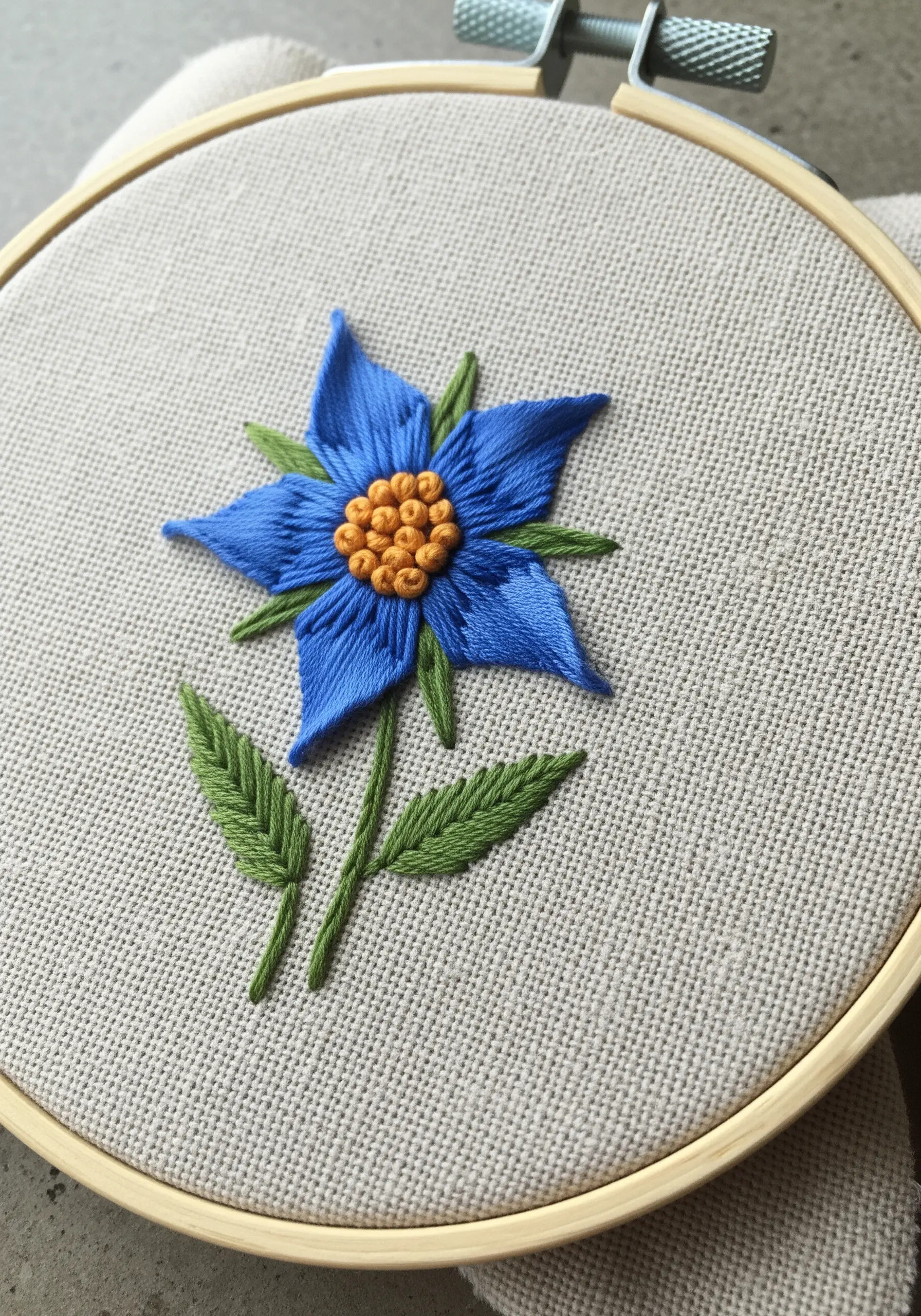 A vibrant blue borage flower with an orange French knot center, stitched on neutral linen.