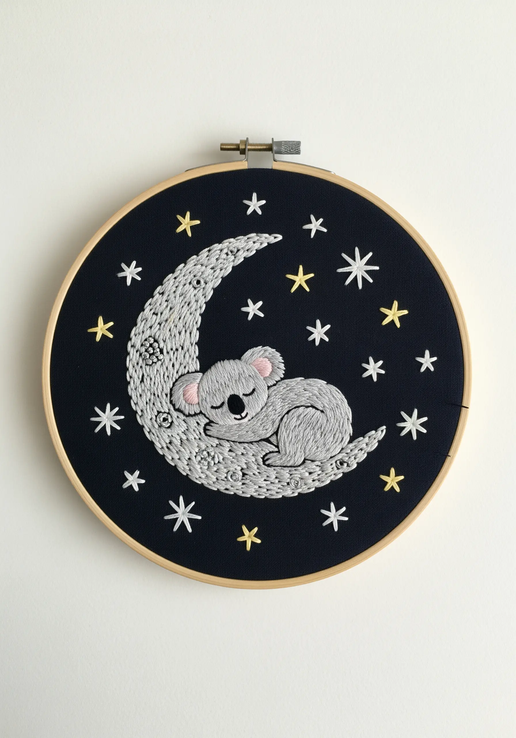 A sleeping koala embroidered on a crescent moon filled with textured stitches, set on dark fabric.