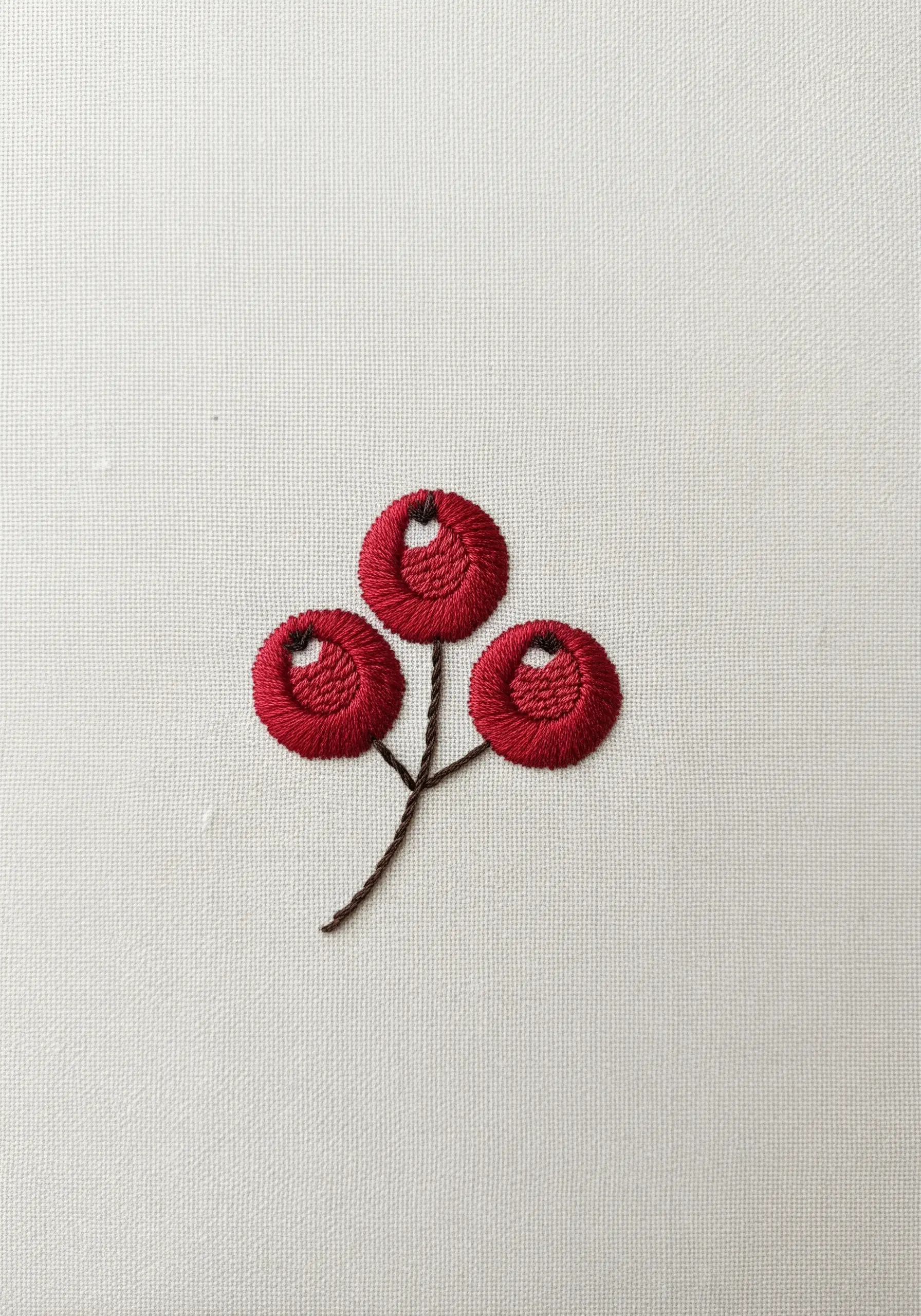 Three red embroidered berries with raised, padded satin stitch on cream fabric.
