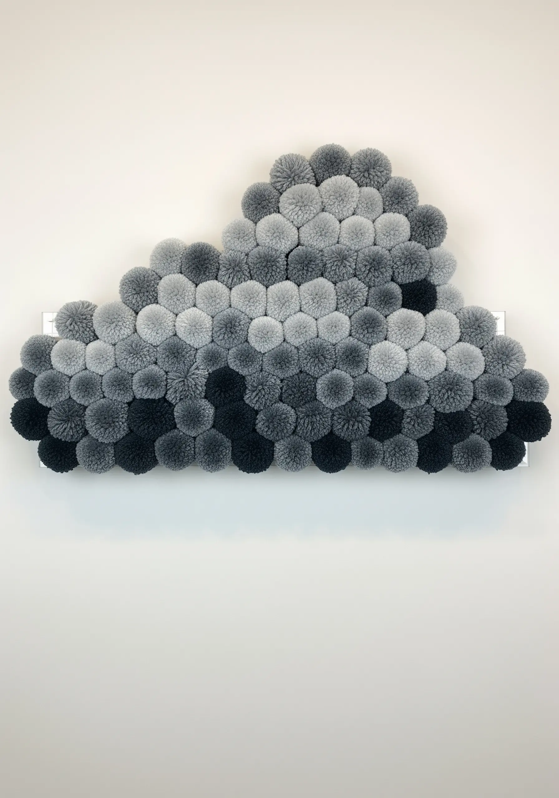 A cloud-shaped wall art piece made of many pom-poms in a grey-scale gradient.