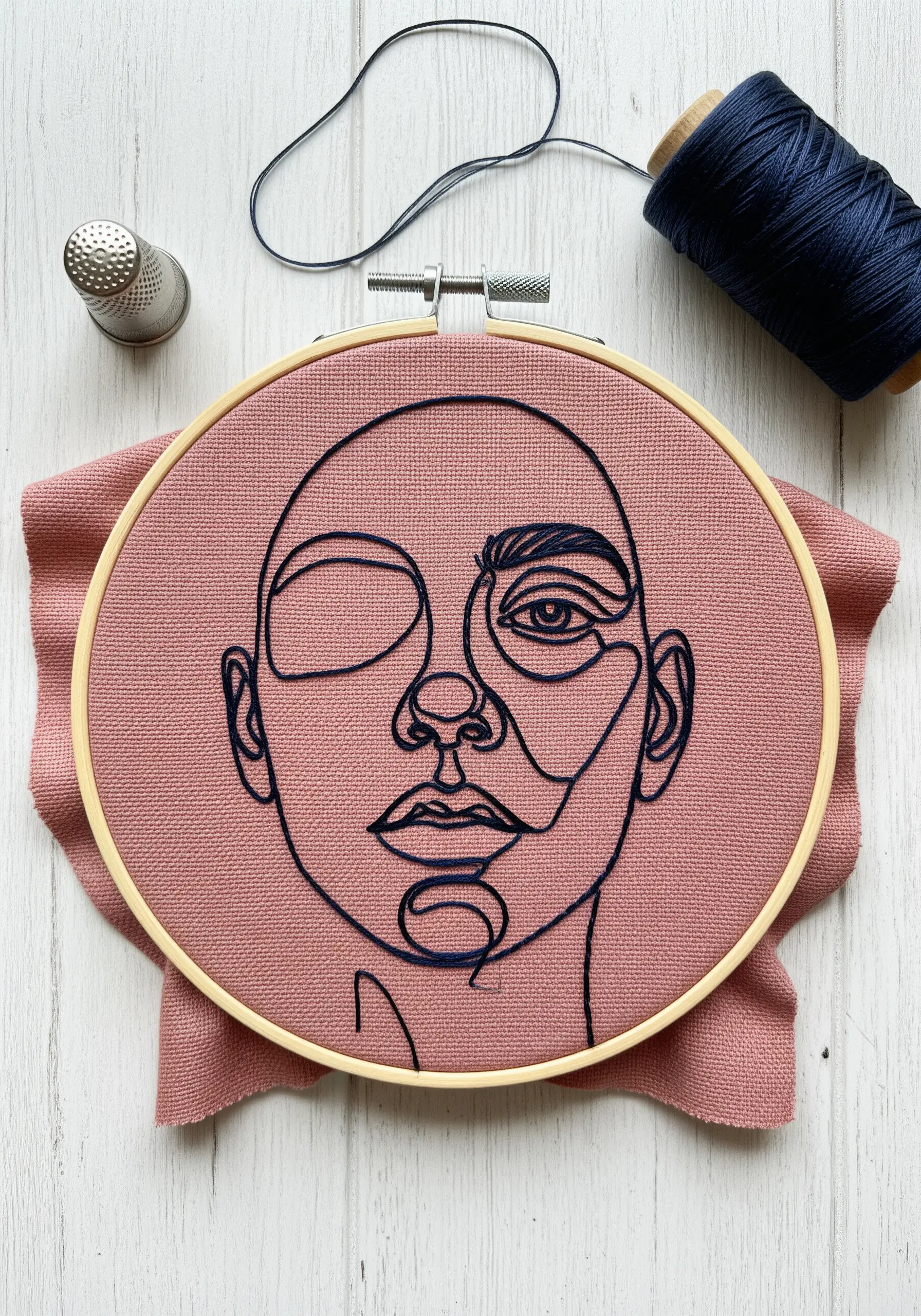 Abstract line art face in dark navy thread on dusty rose fabric with varied line weights.