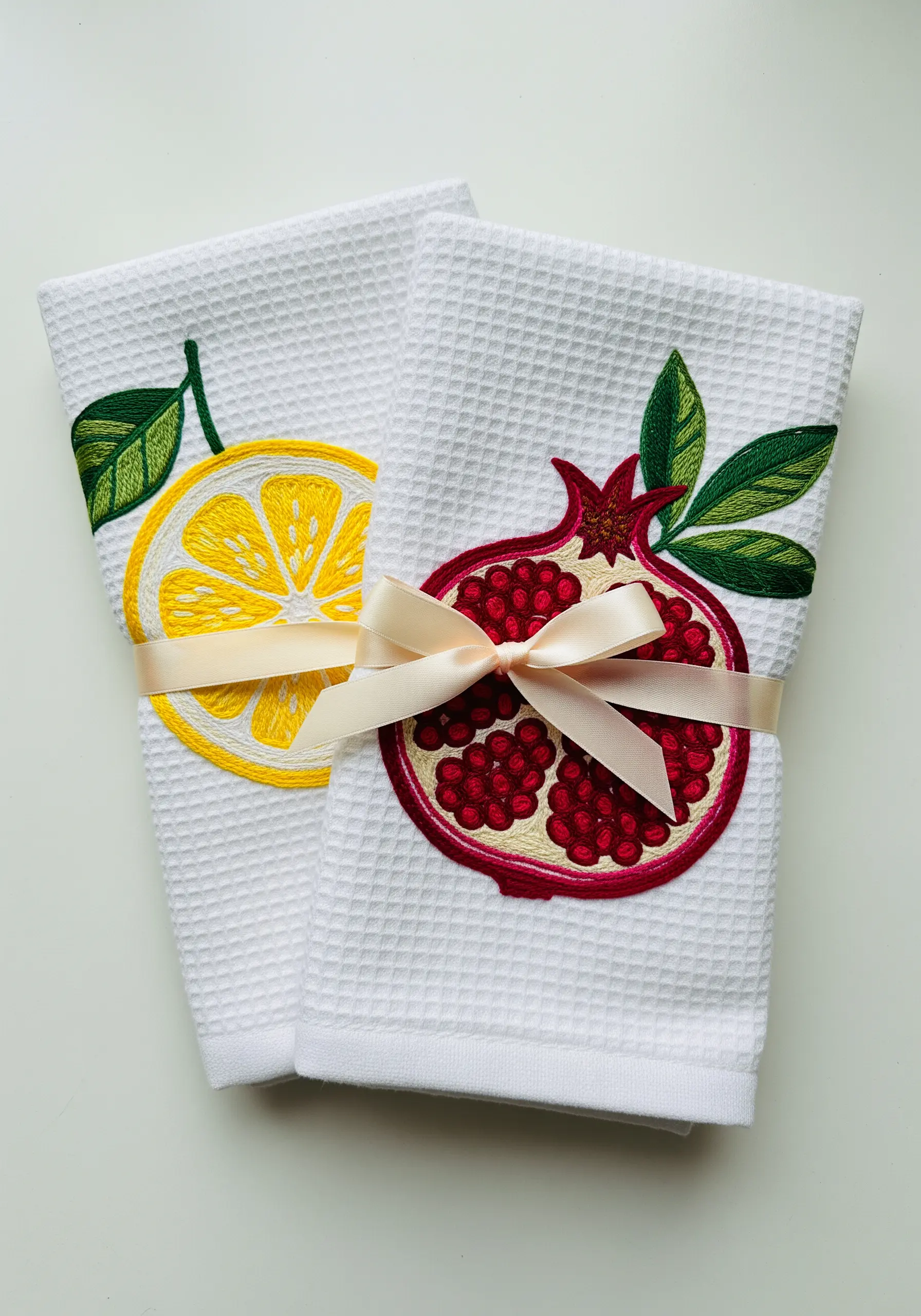 Two white waffle-weave kitchen towels, one embroidered with a lemon slice and one with a pomegranate.
