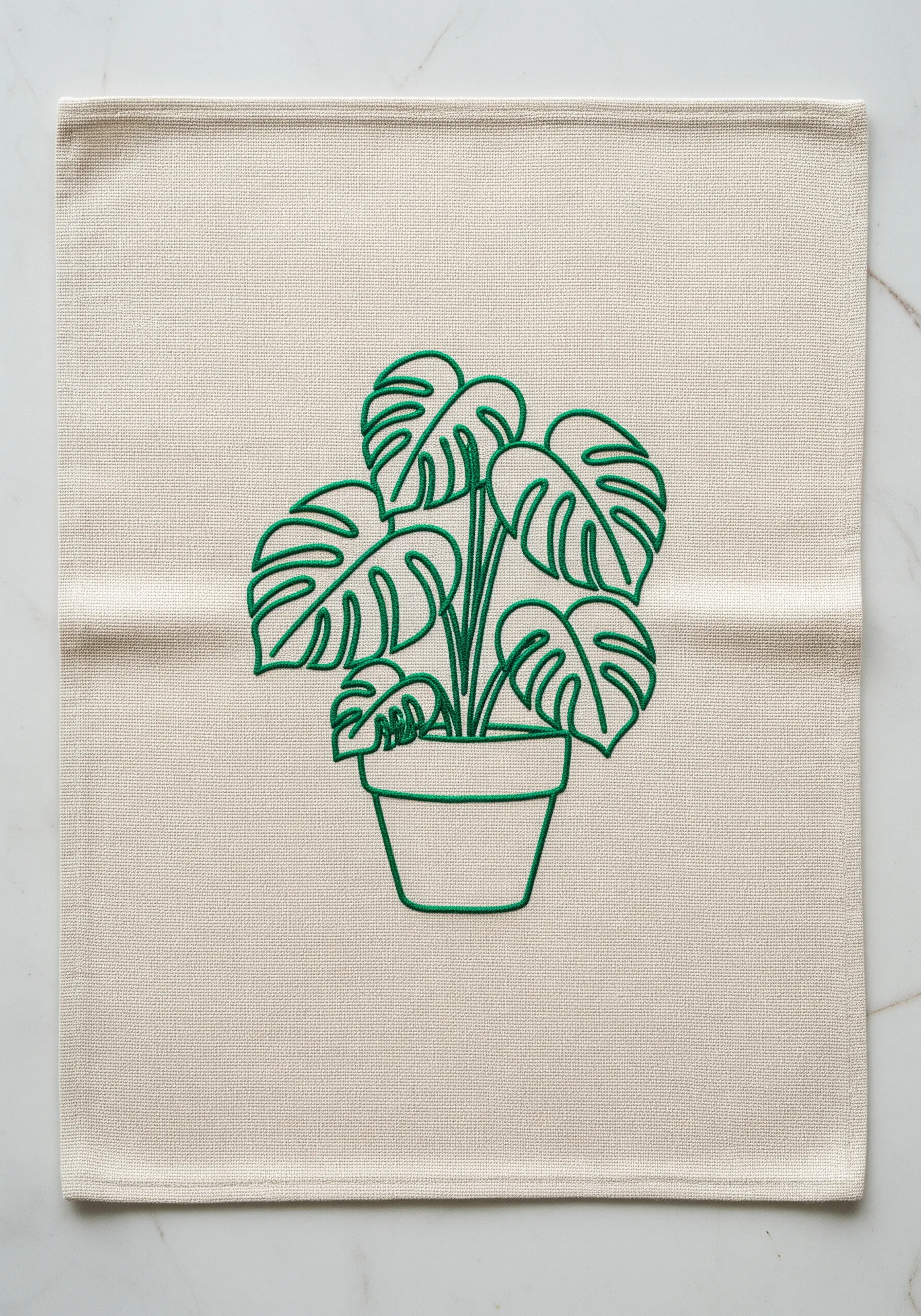 A bold green line-art embroidery of a potted monstera plant on a neutral linen placemat.