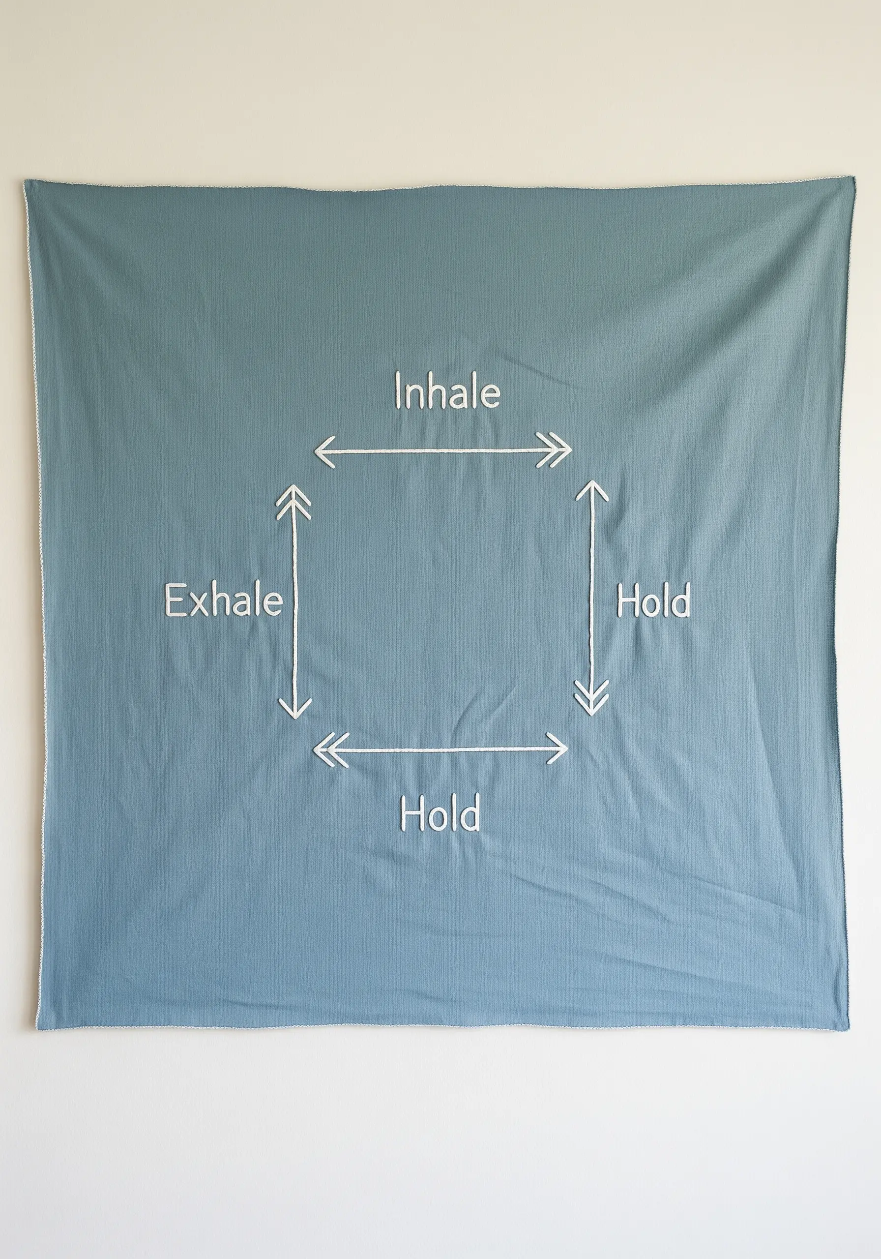 A blue wall tapestry embroidered with the words 'Inhale, Hold, Exhale, Hold' in a box formation.