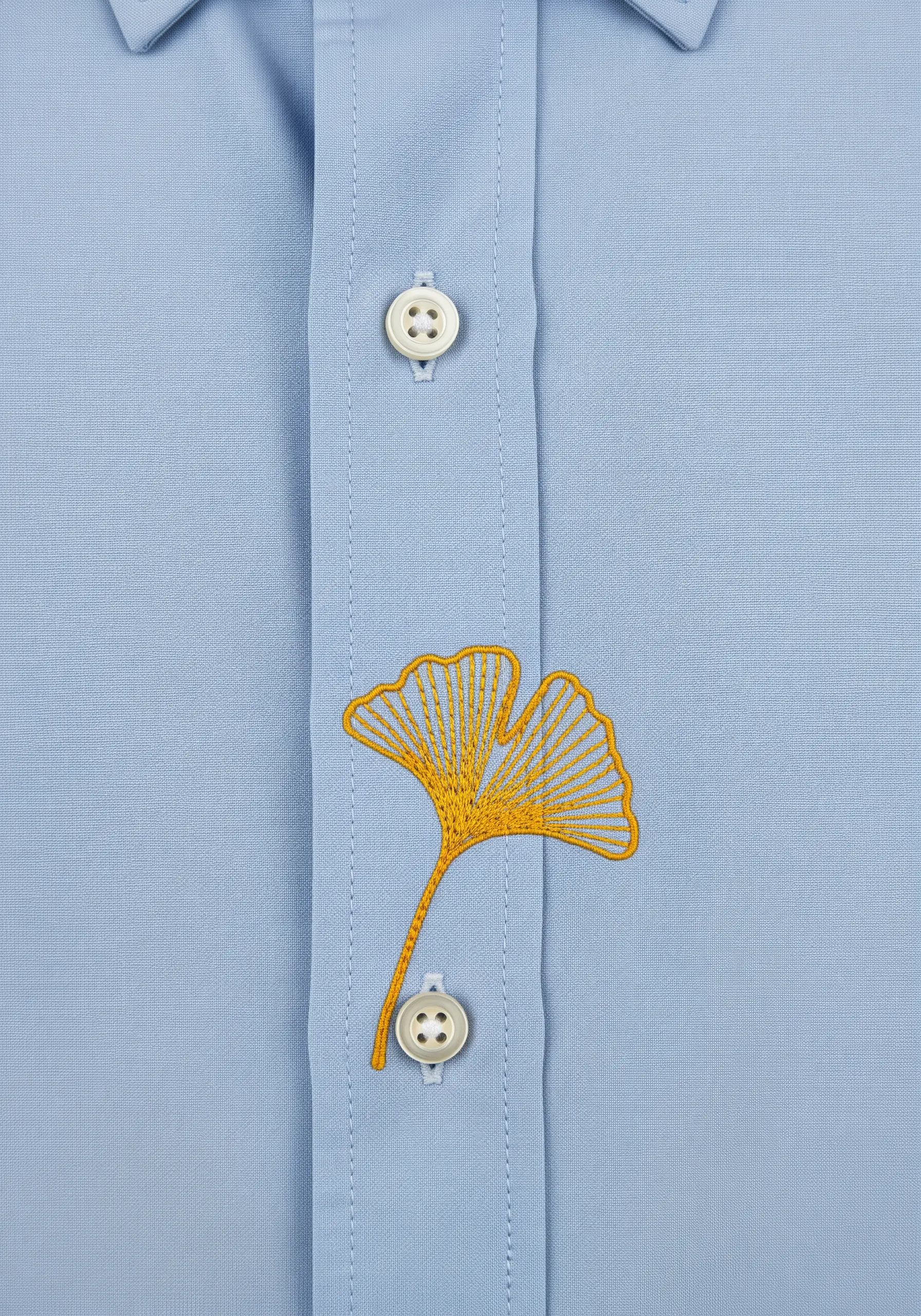 A yellow ginkgo leaf embroidered on a light blue shirt, positioned between two buttons.