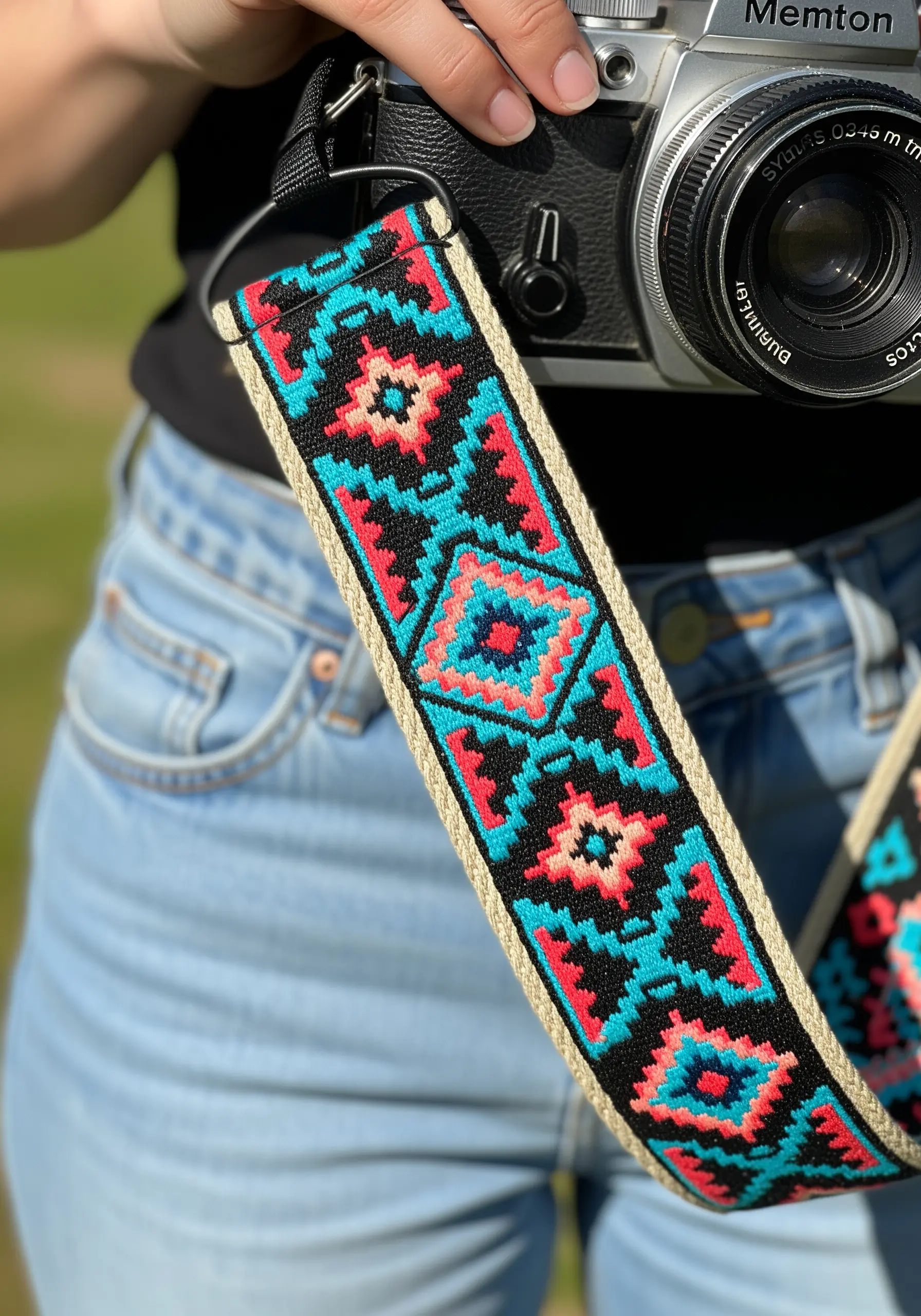 A camera strap with a geometric, woven-look embroidery pattern in teal, pink, and black.