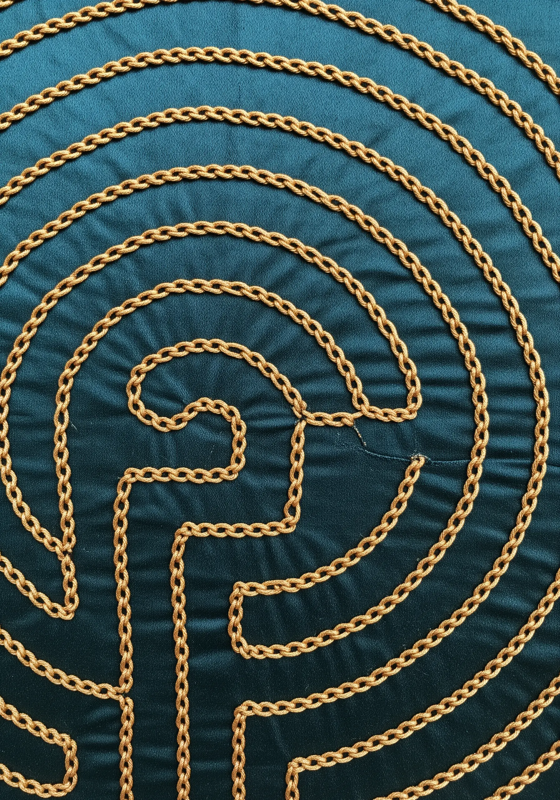 A close-up of a labyrinth pattern created with gold chain stitch on dark teal fabric.