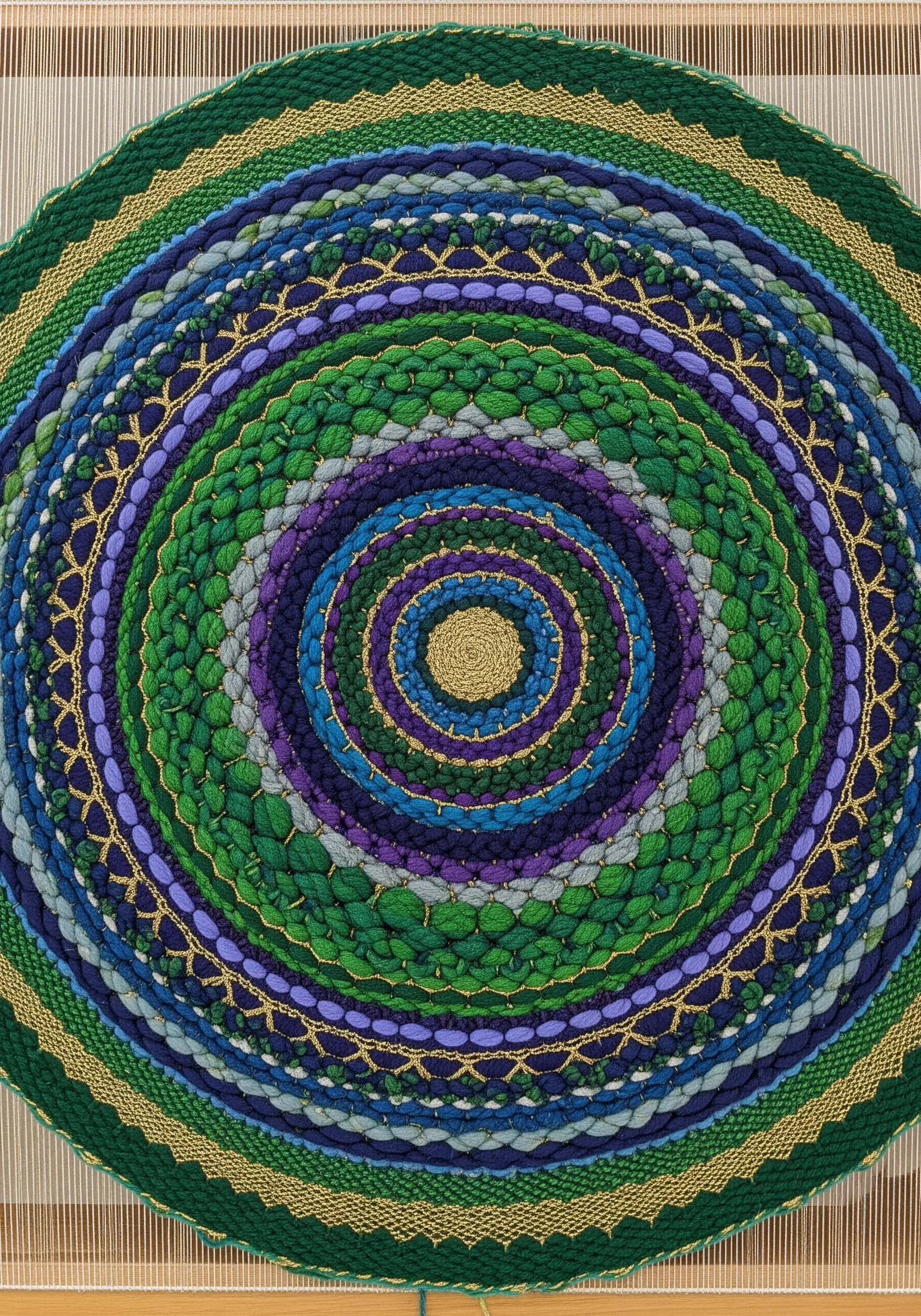 A close-up of a circular woven mandala with concentric rings of green, blue, and purple yarn.