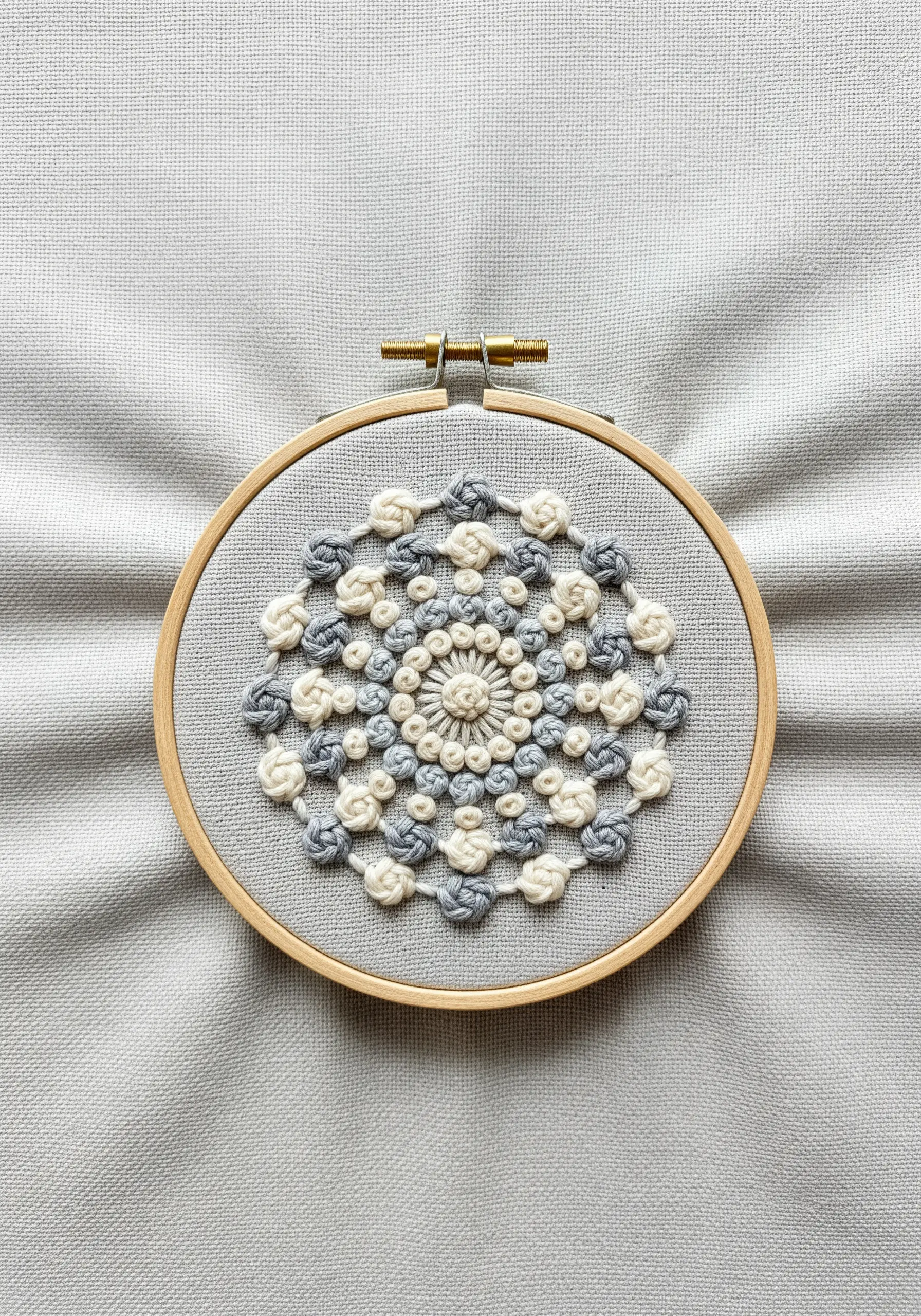 A highly textured mandala made of grey and cream knots on grey fabric.