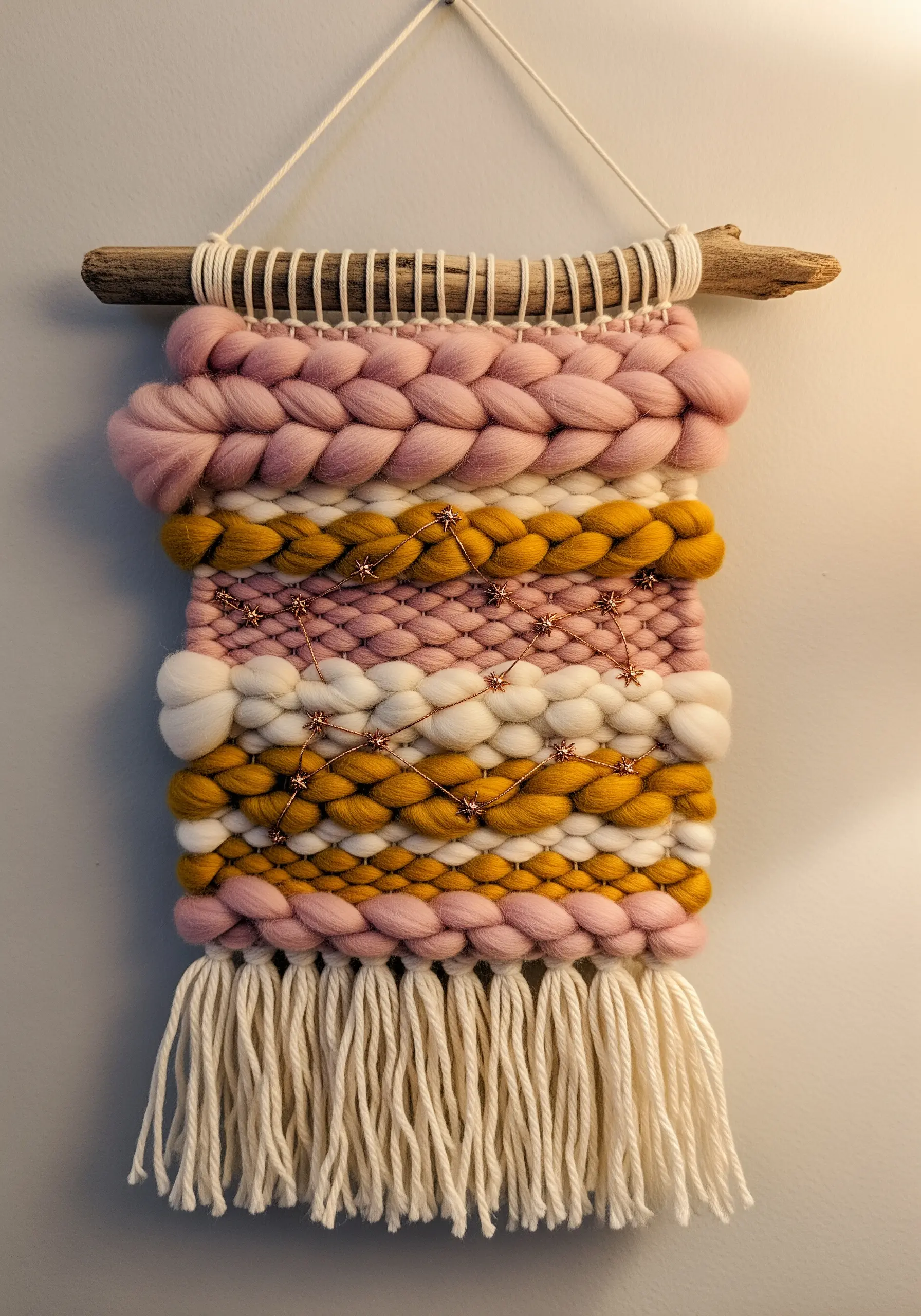 A woven wall hanging with thick pink and yellow yarn, featuring embroidered copper constellations.