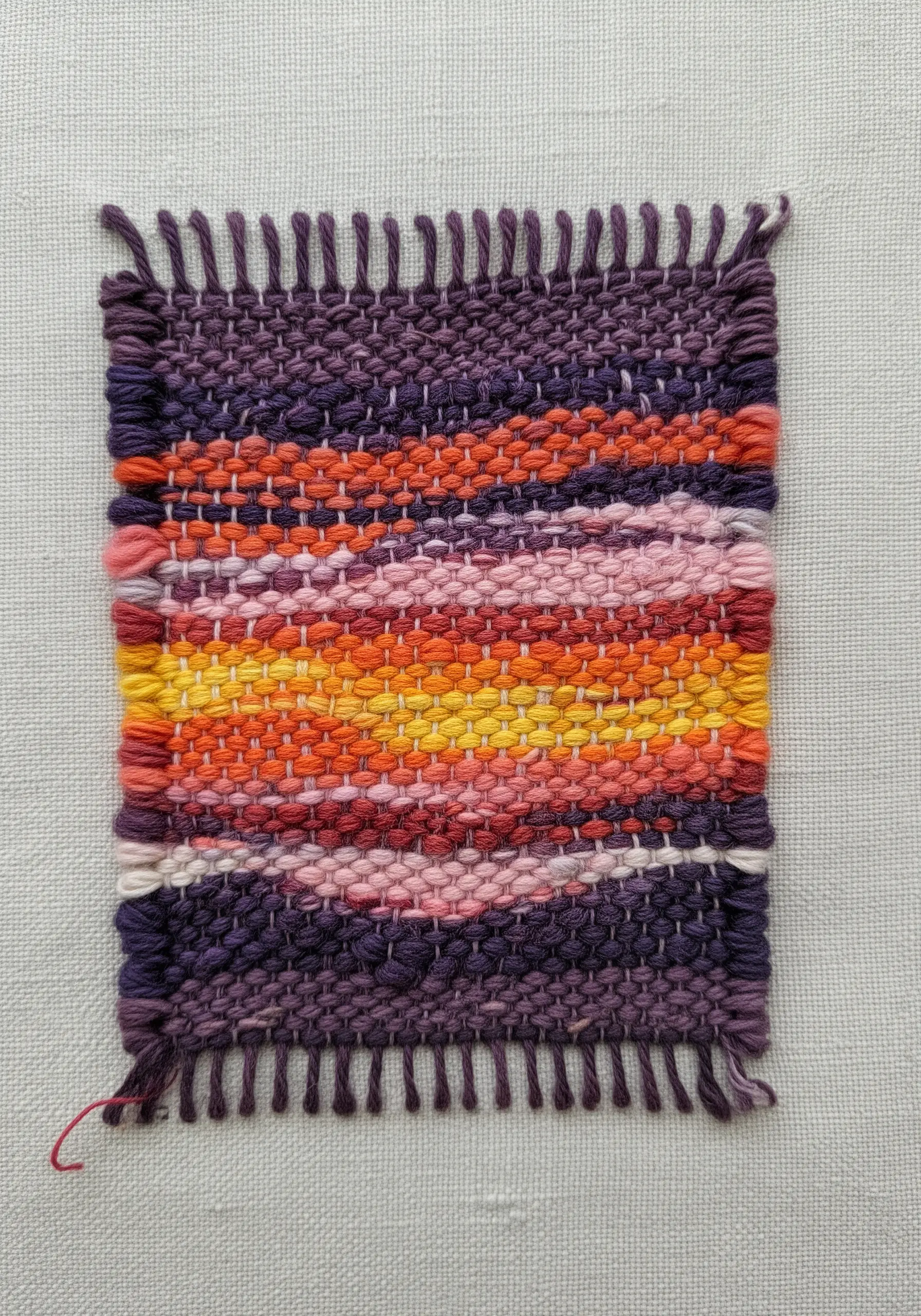 A small, colorful woven patch with a sunset-like gradient of purples, pinks, oranges, and yellows.