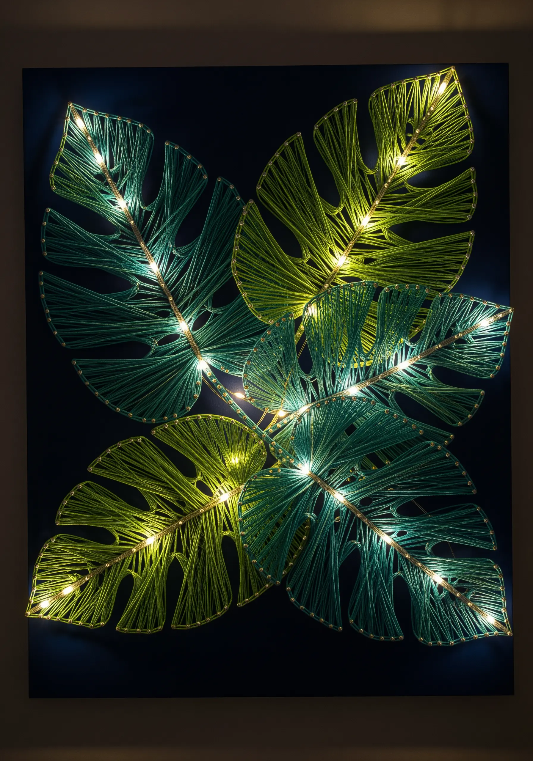 String art of four Monstera leaves on a dark background, with small LED lights illuminating the veins.