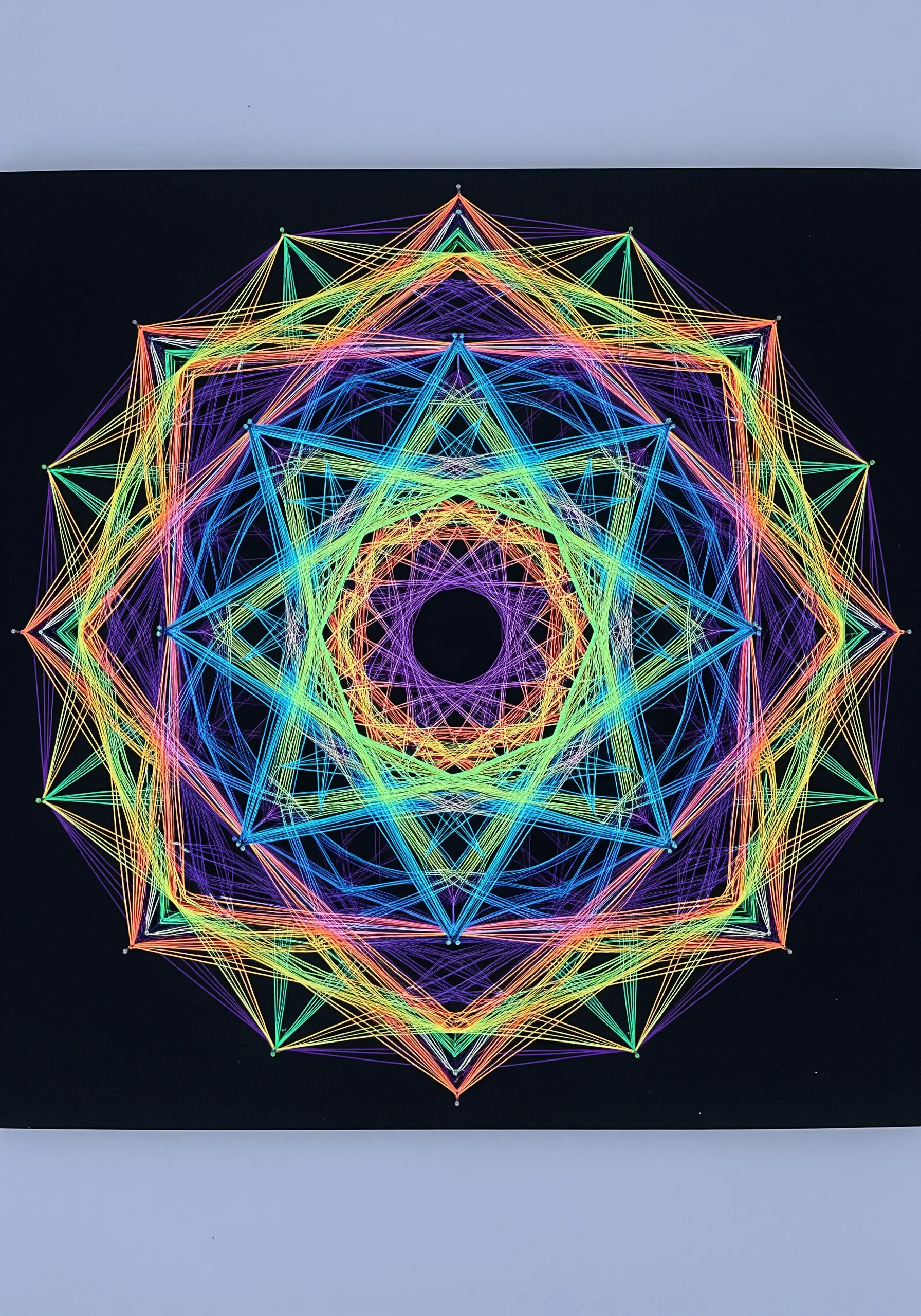 A complex geometric mandala made with layered rainbow neon threads on black canvas.