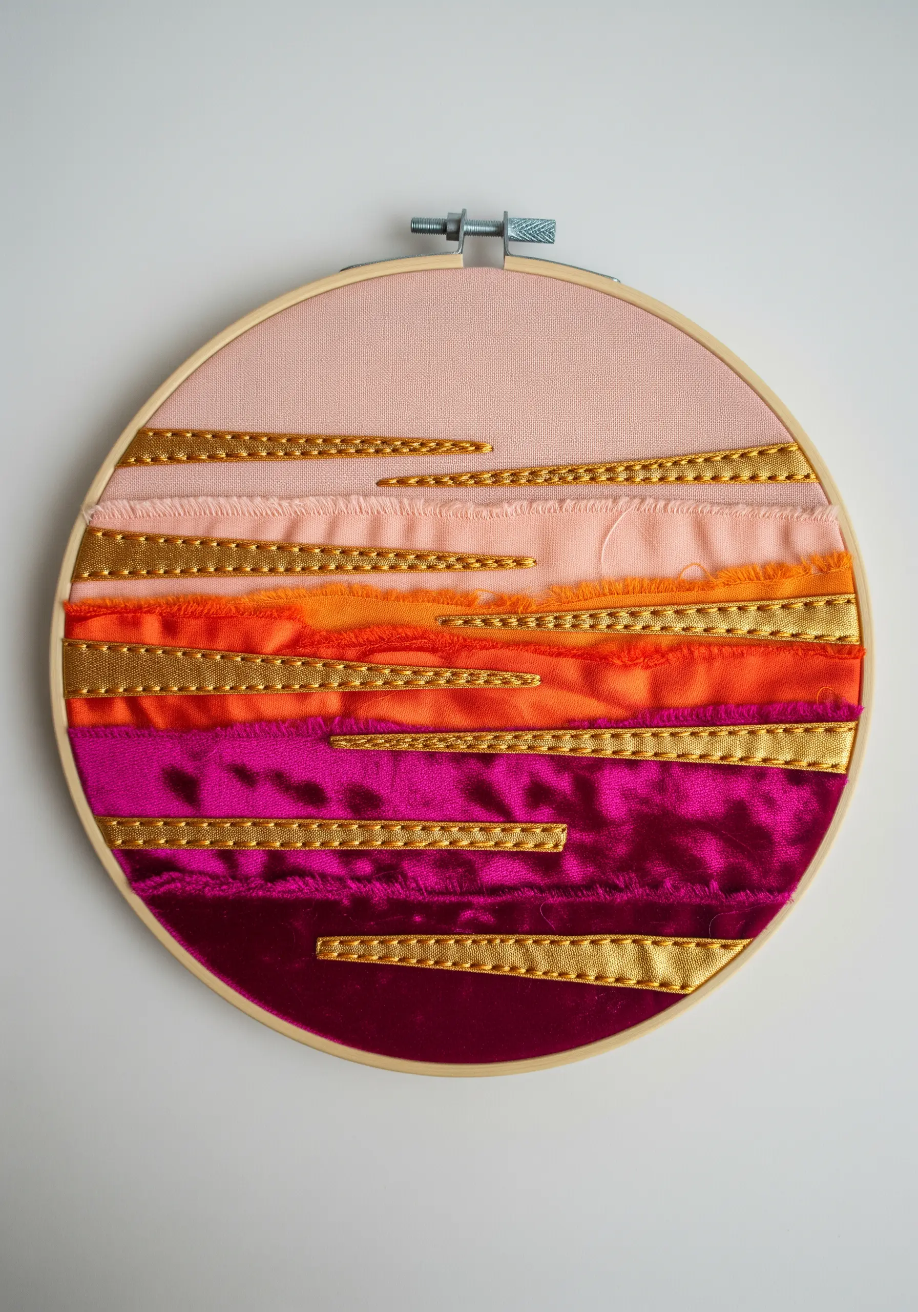 Abstract sunset embroidery with layered fabric strips and woven gold ribbon.