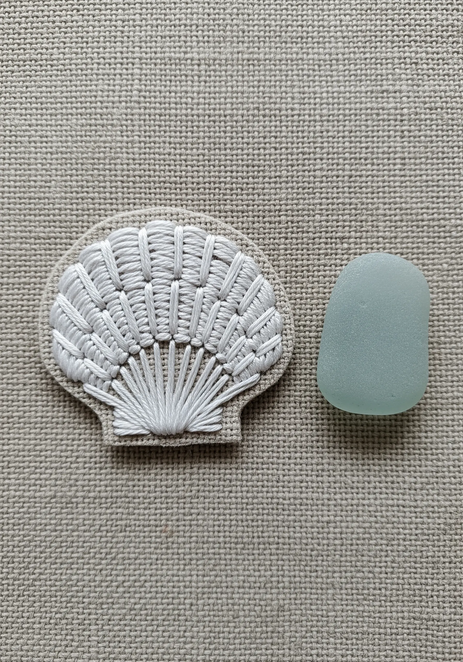 An embroidered seashell patch using white woven stitches to create raised, linear texture.