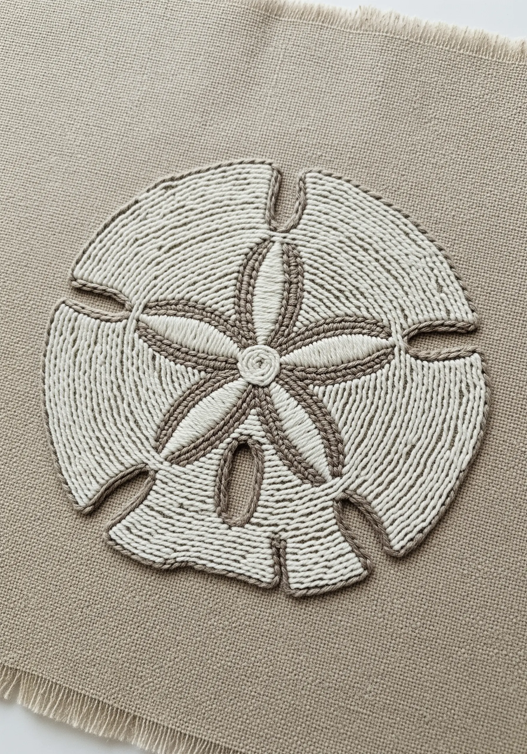 A sand dollar embroidered with dense, textured laid work in cream and taupe threads.
