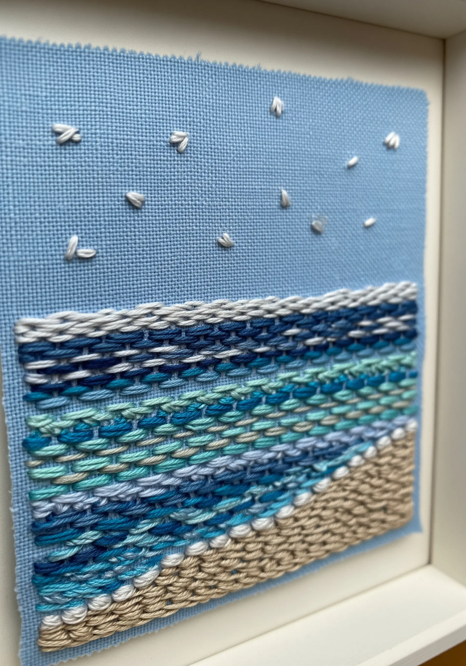 A woven embroidery seascape with textured blue waves and a sandy shore.