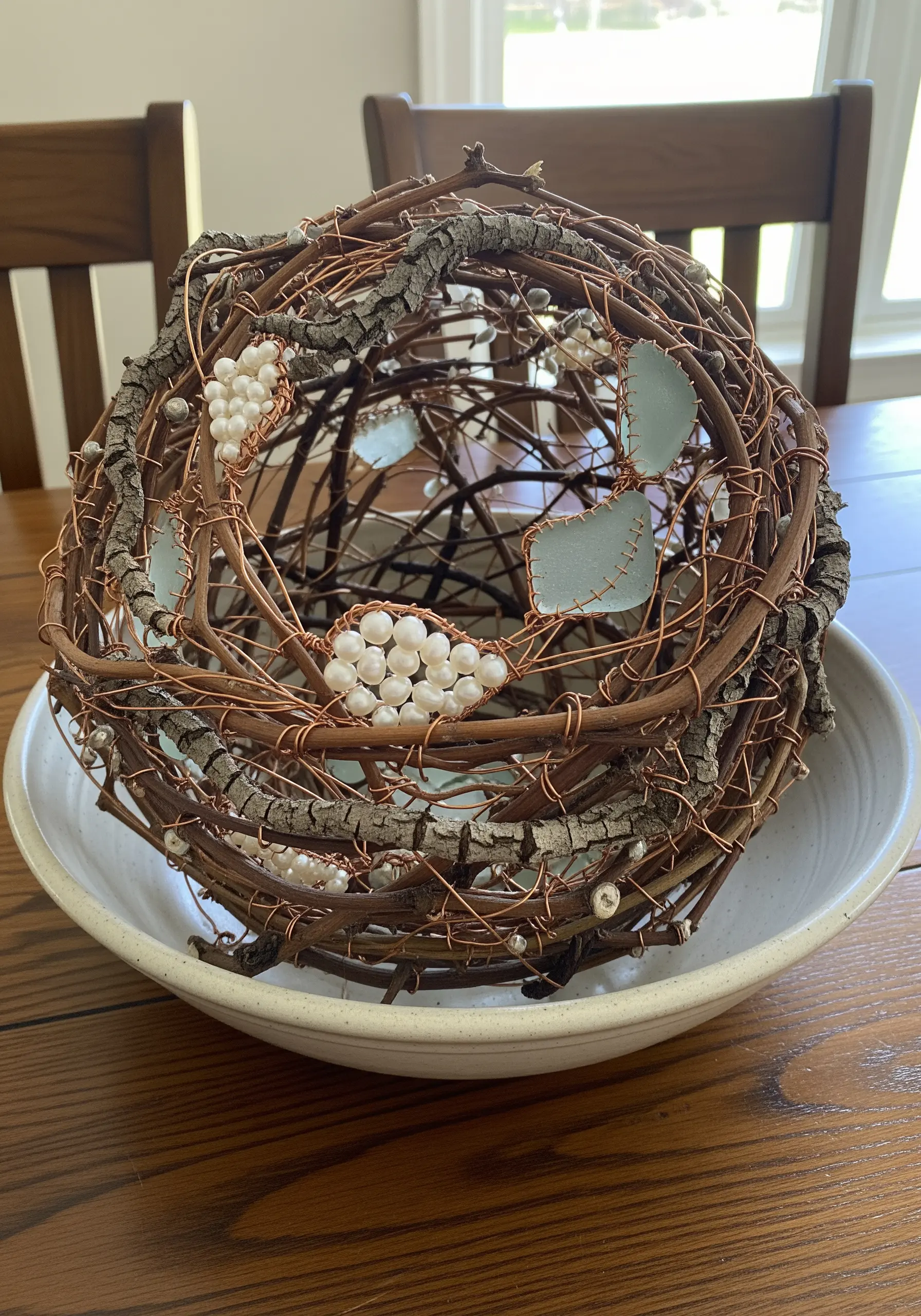 A spherical sculpture made of woven twigs, copper wire, sea glass, and clusters of white pearls.