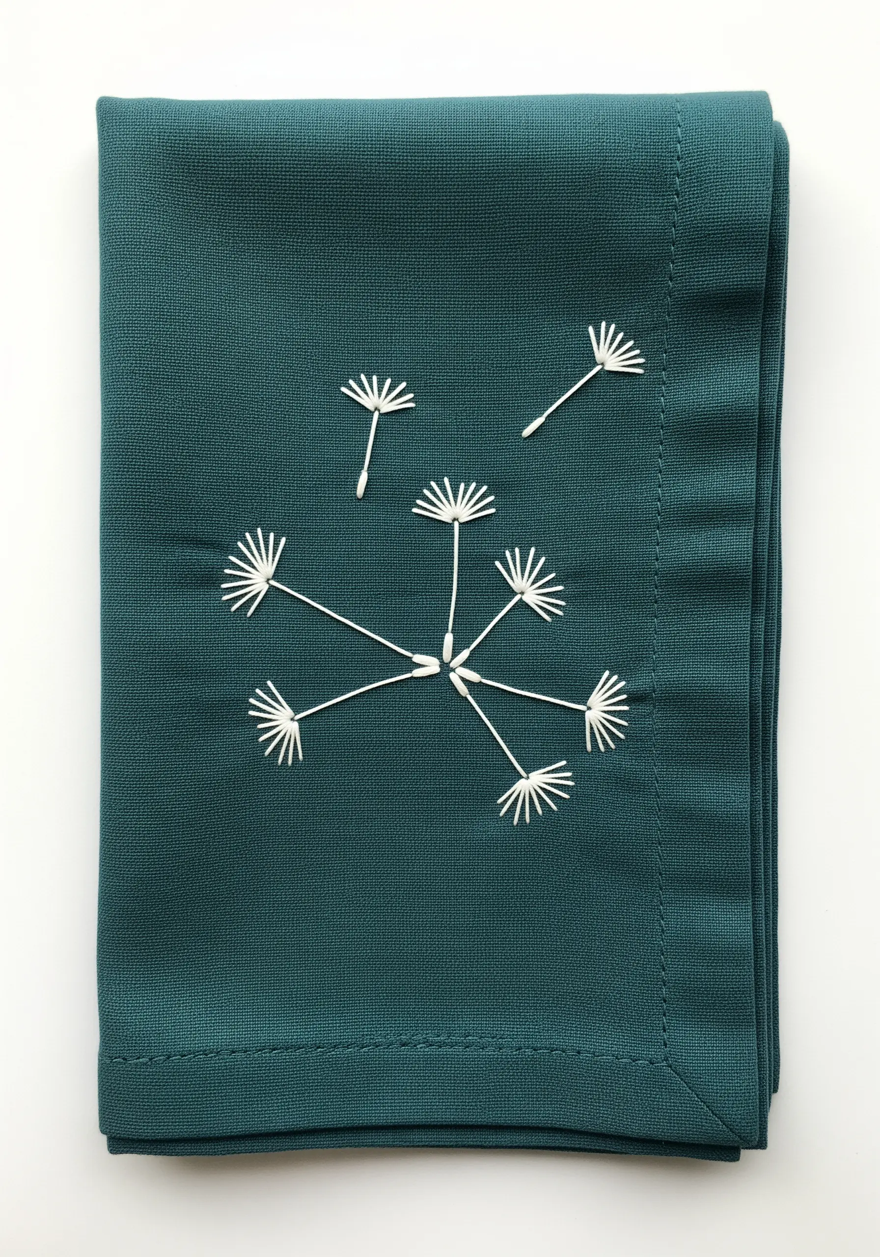 Delicate dandelion seeds embroidered in white thread on a dark teal napkin.