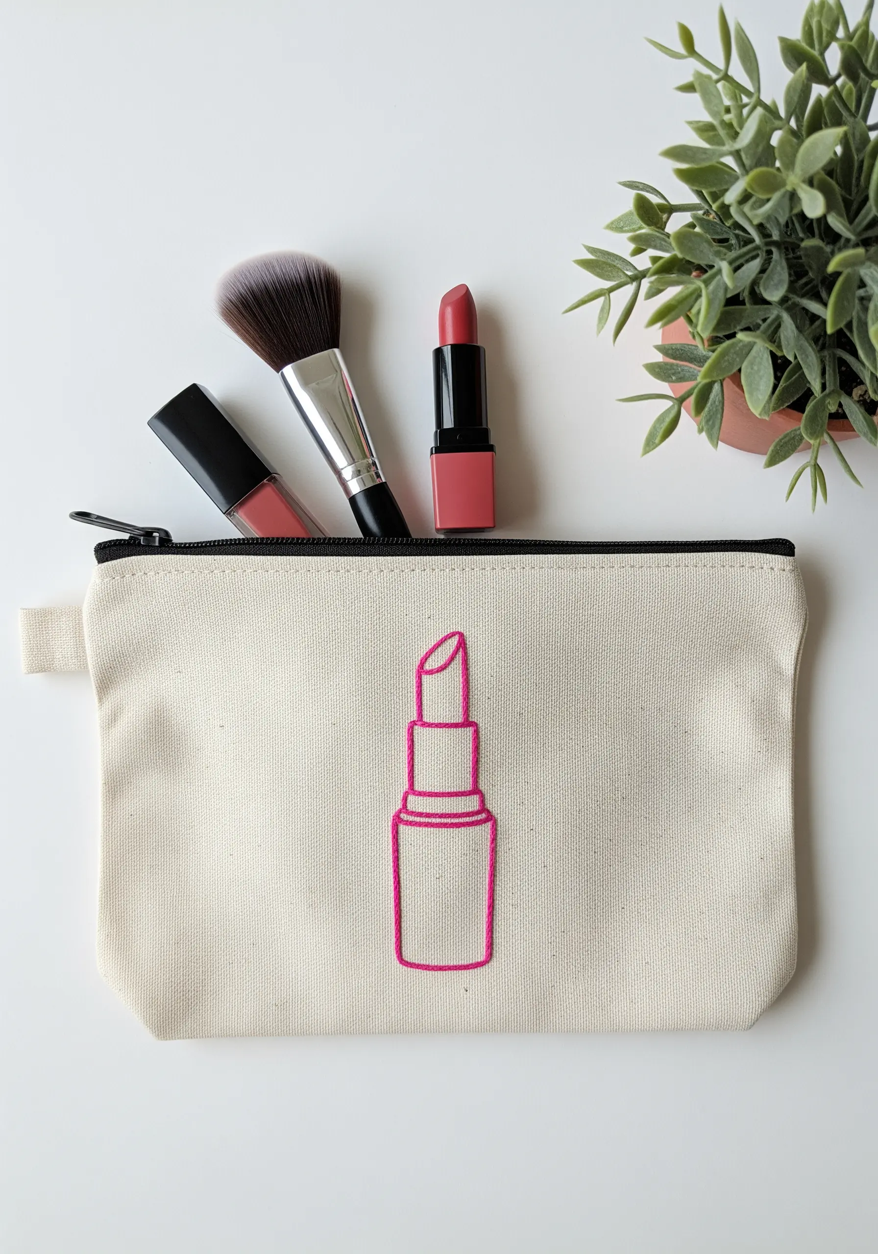 A canvas makeup pouch embroidered with the bright pink outline of a tube of lipstick.