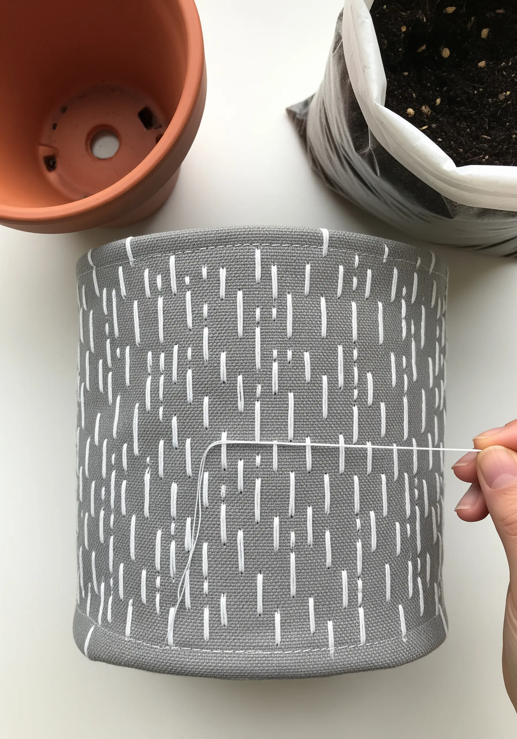 A grey fabric plant pot cover being decorated with a pattern of short, white vertical stitches.