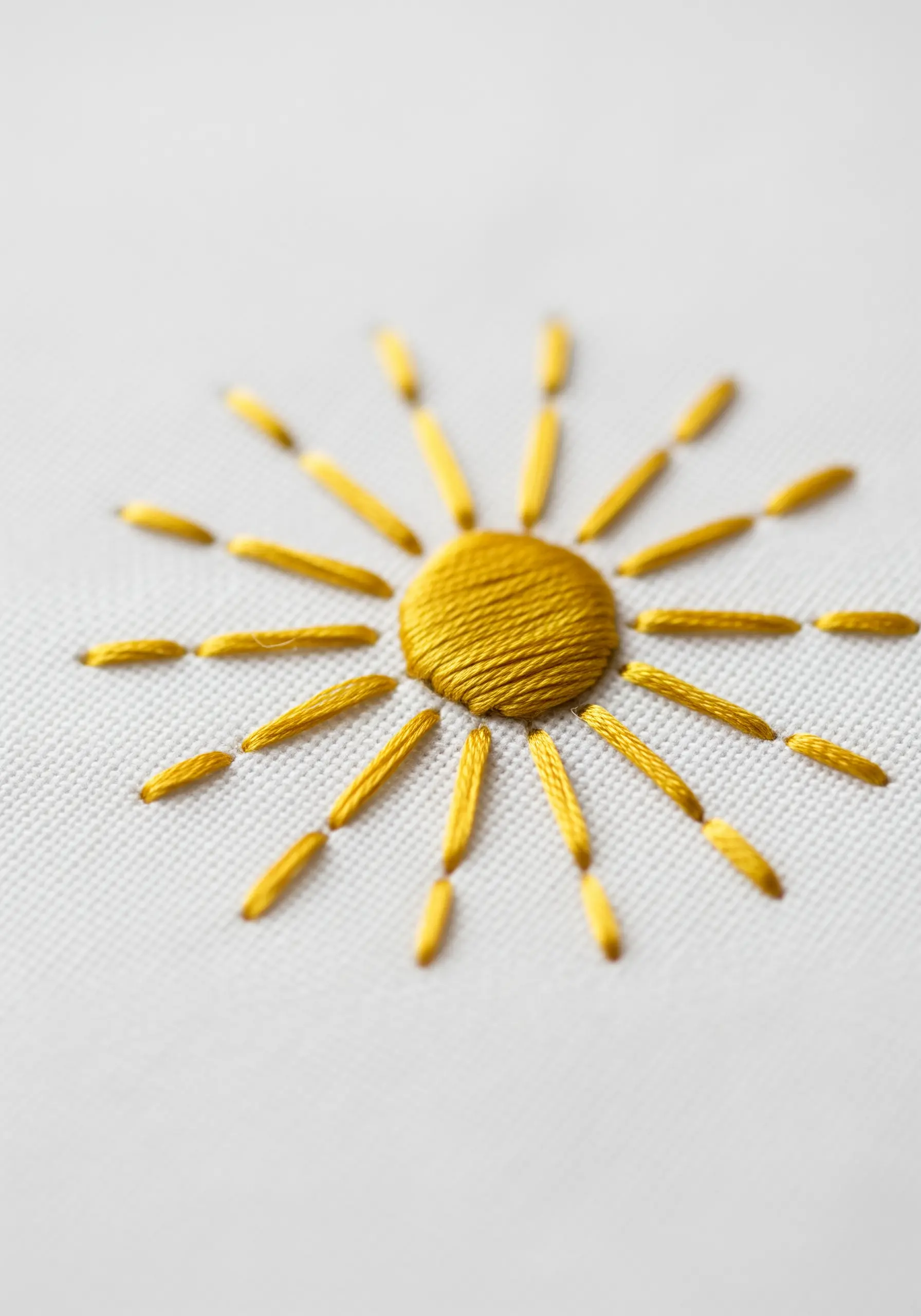 A close-up of a minimalist sun embroidered with a golden satin stitch circle and straight stitch rays.