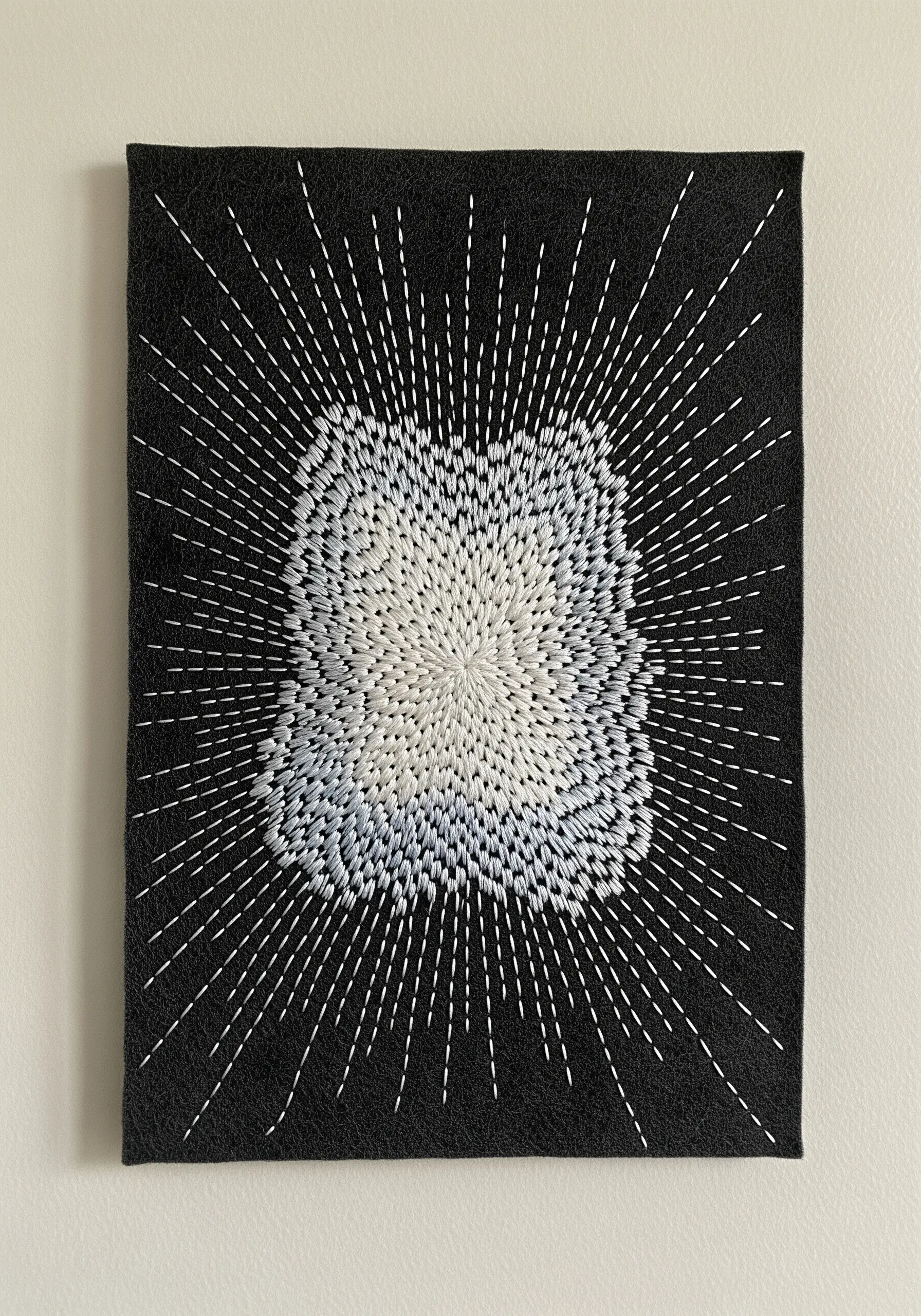 White and pale blue seed stitches radiate outward on black felt fabric, creating a starburst effect.