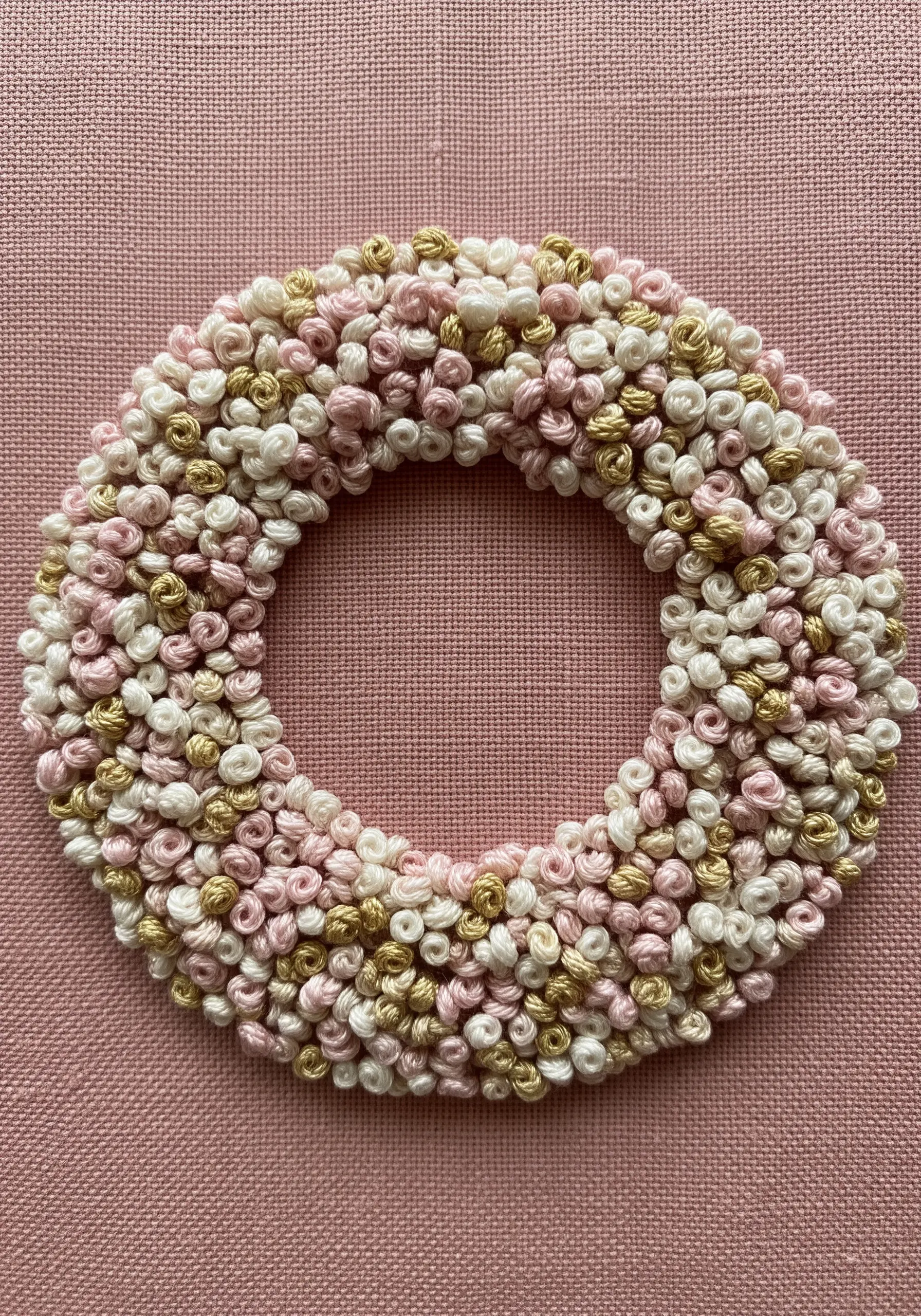 A dense, textured wreath made entirely of pink, white, and gold French knots on pink fabric.