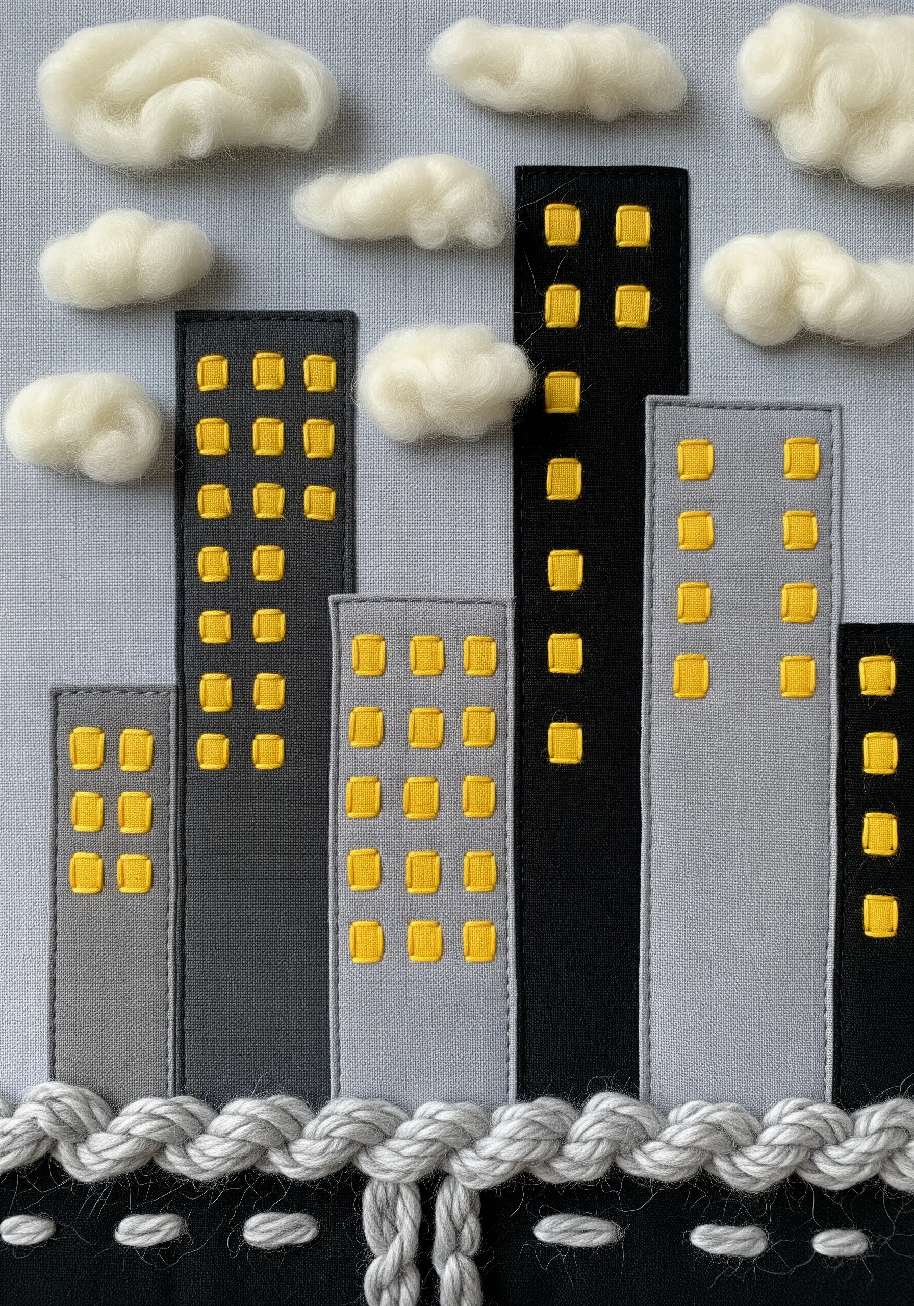 A fabric appliqué cityscape with gray buildings, yellow windows, and soft, needle-felted white clouds.
