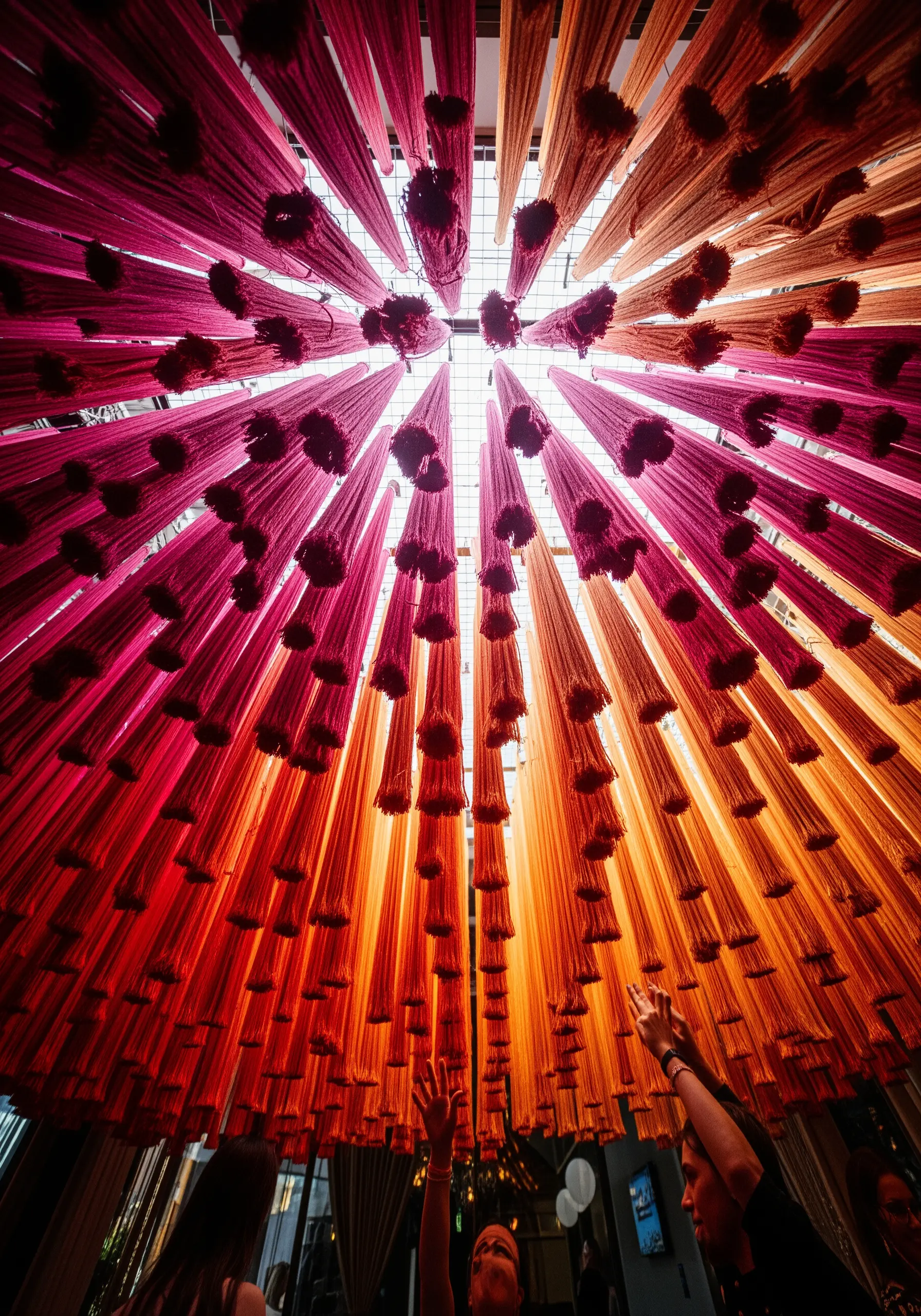 Looking up at a ceiling installation of thousands of hanging pink, red, and orange yarn tassels.