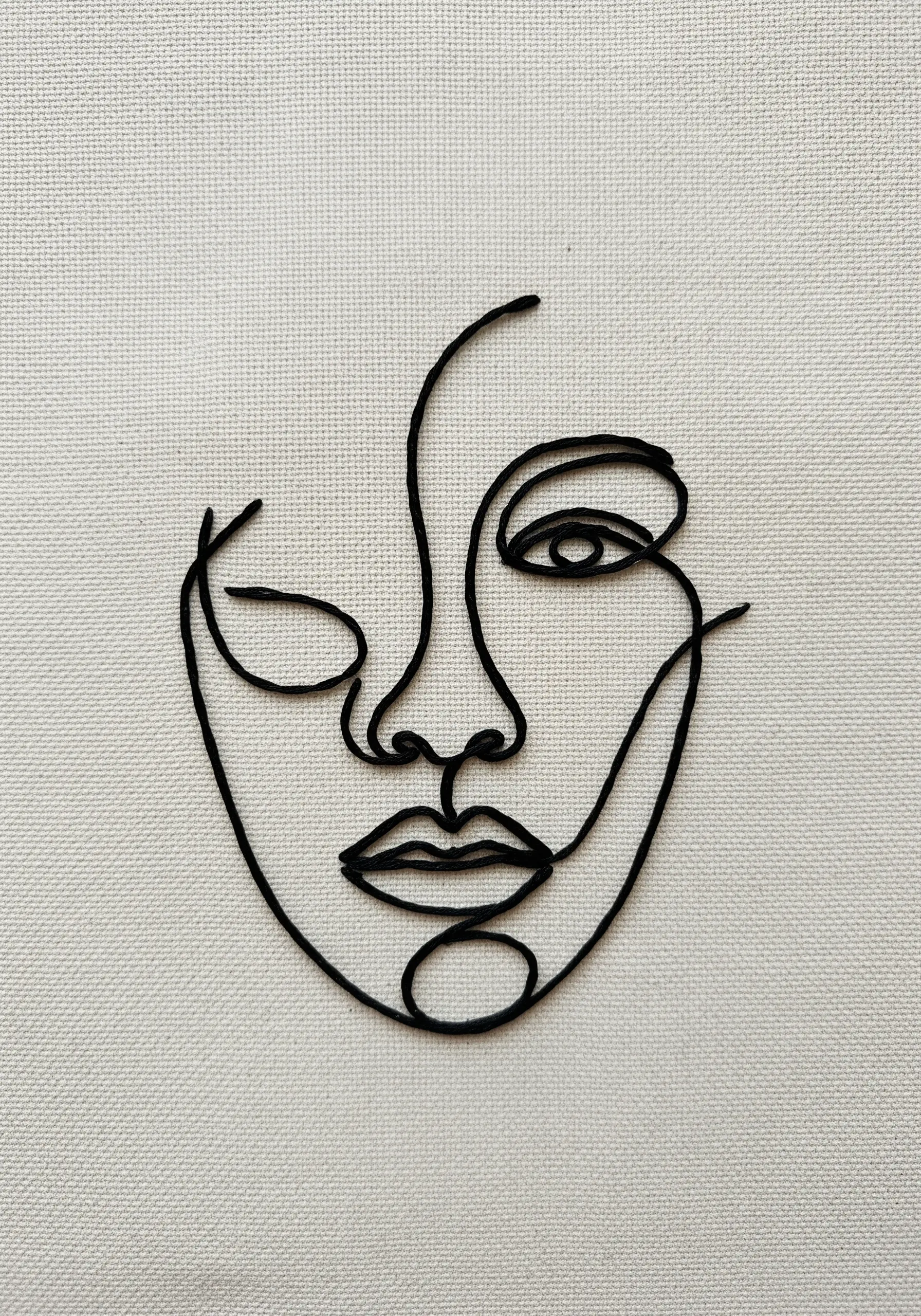 A minimalist abstract face embroidered in a single, continuous black line on off-white fabric.