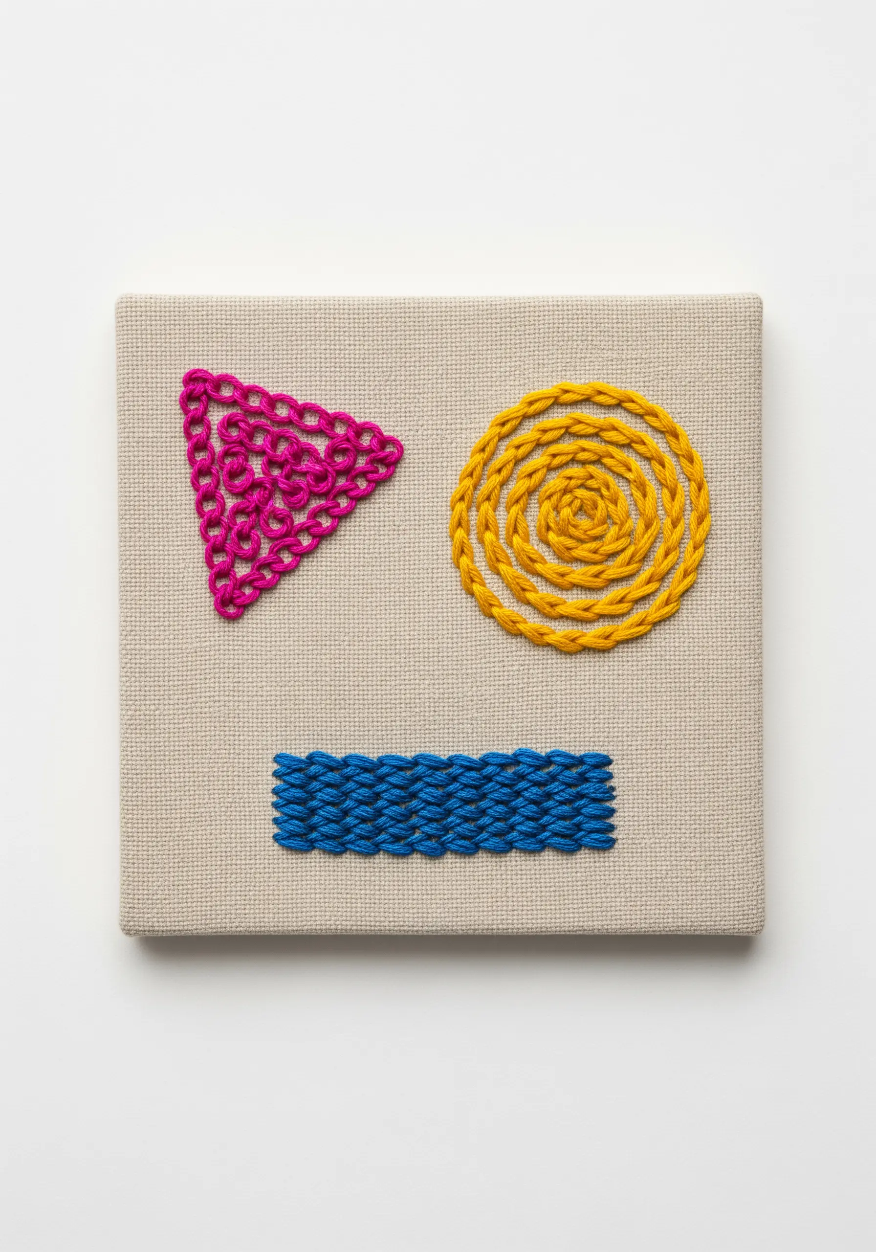 Abstract shapes on canvas: a pink triangle in Pekinese stitch, a yellow circle in a coil, and a blue rectangle.