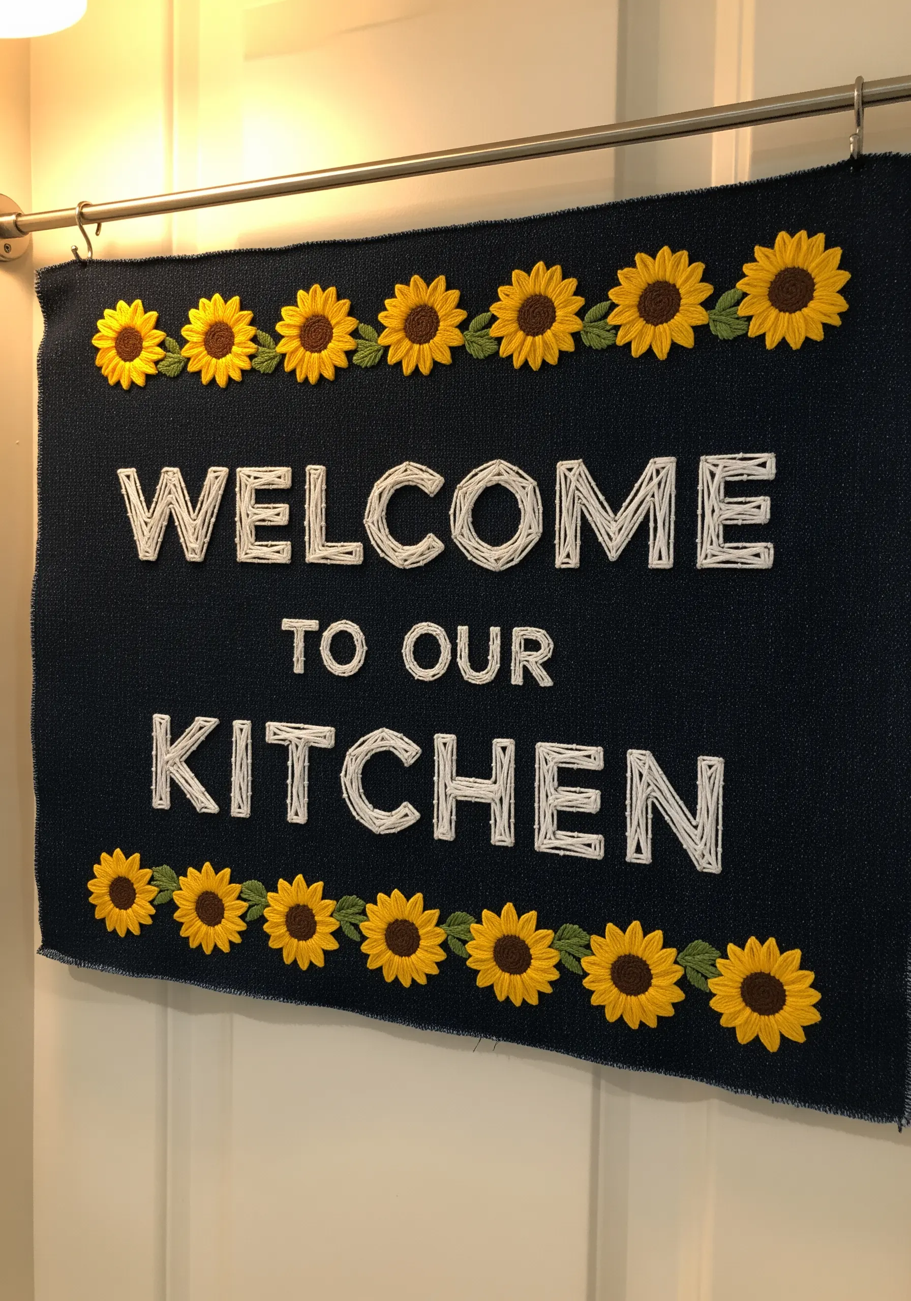 A kitchen welcome sign with white text bordered by 3D embroidered sunflowers on dark fabric.