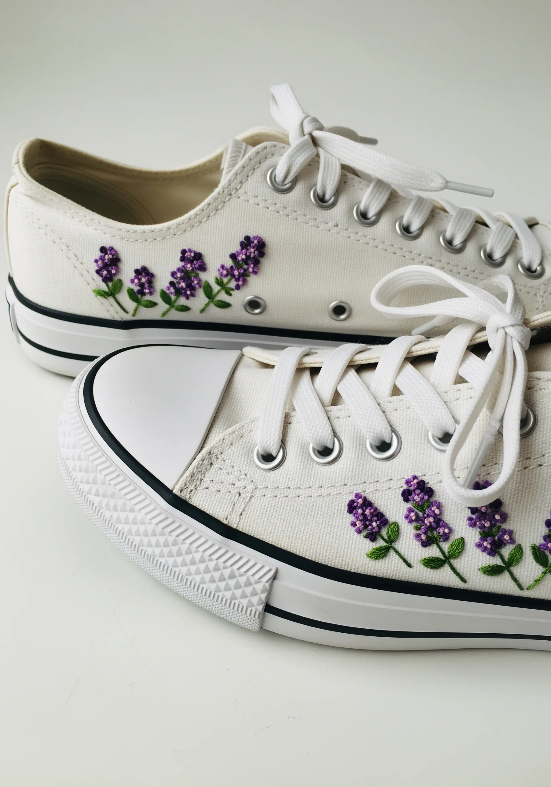 Small clusters of purple lilac flowers embroidered along the sides of white canvas sneakers.