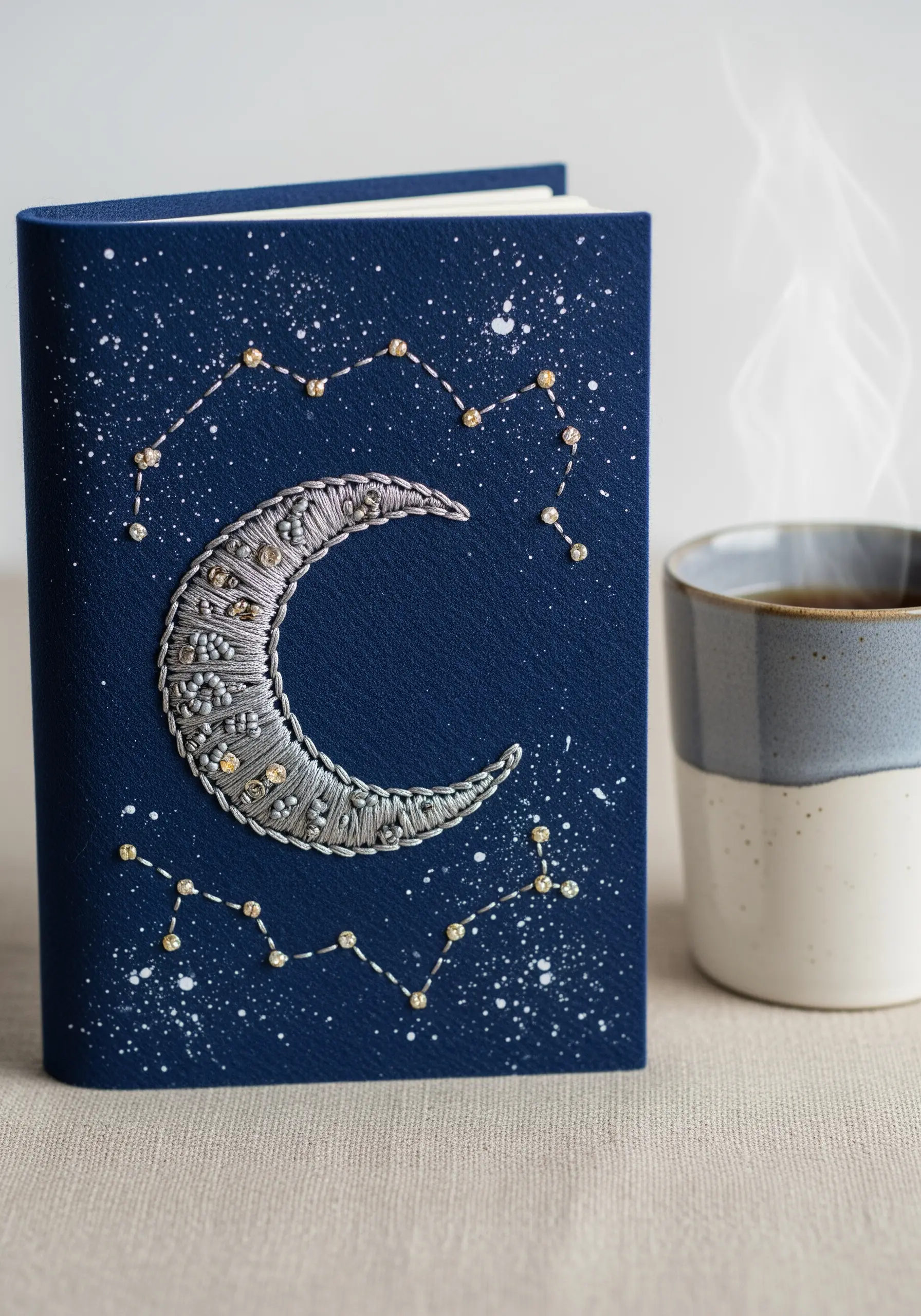 A crescent moon embroidered with silver thread and beads on a dark blue journal cover.