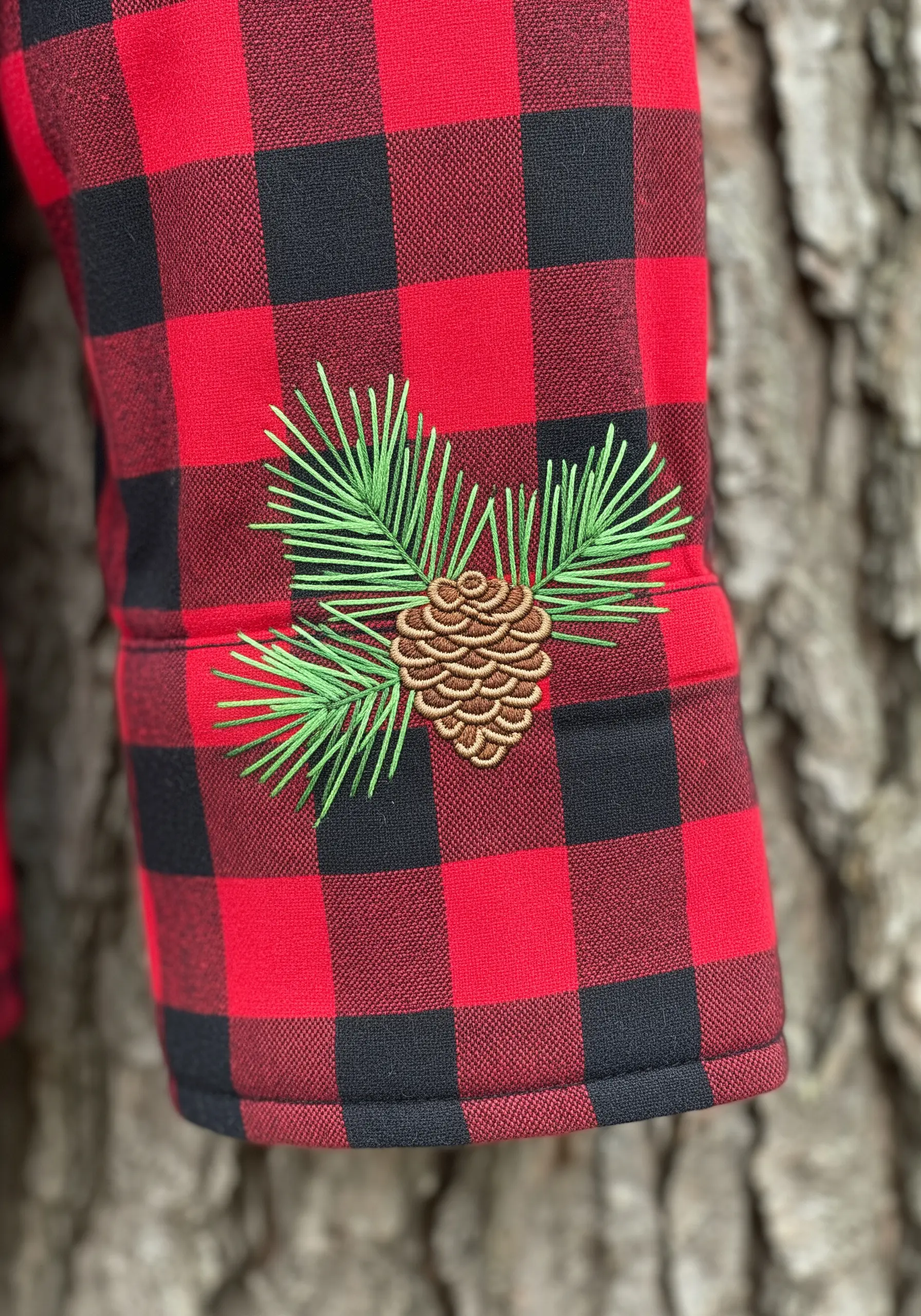 An embroidered pinecone with green needles stitched on the cuff of a red and black plaid shirt.