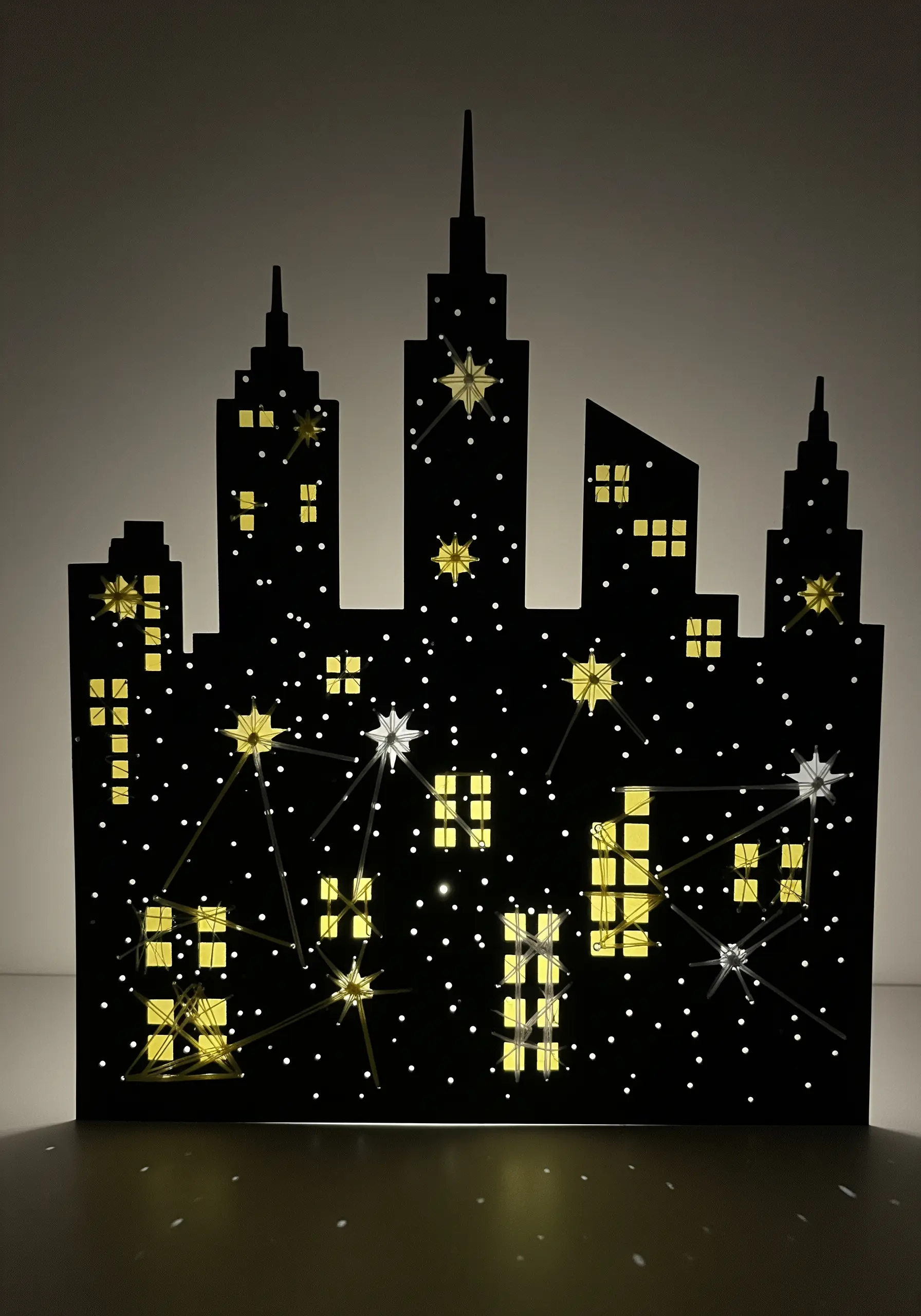 A black paper cityscape silhouette with stitched stars and constellations backlit by yellow lights.